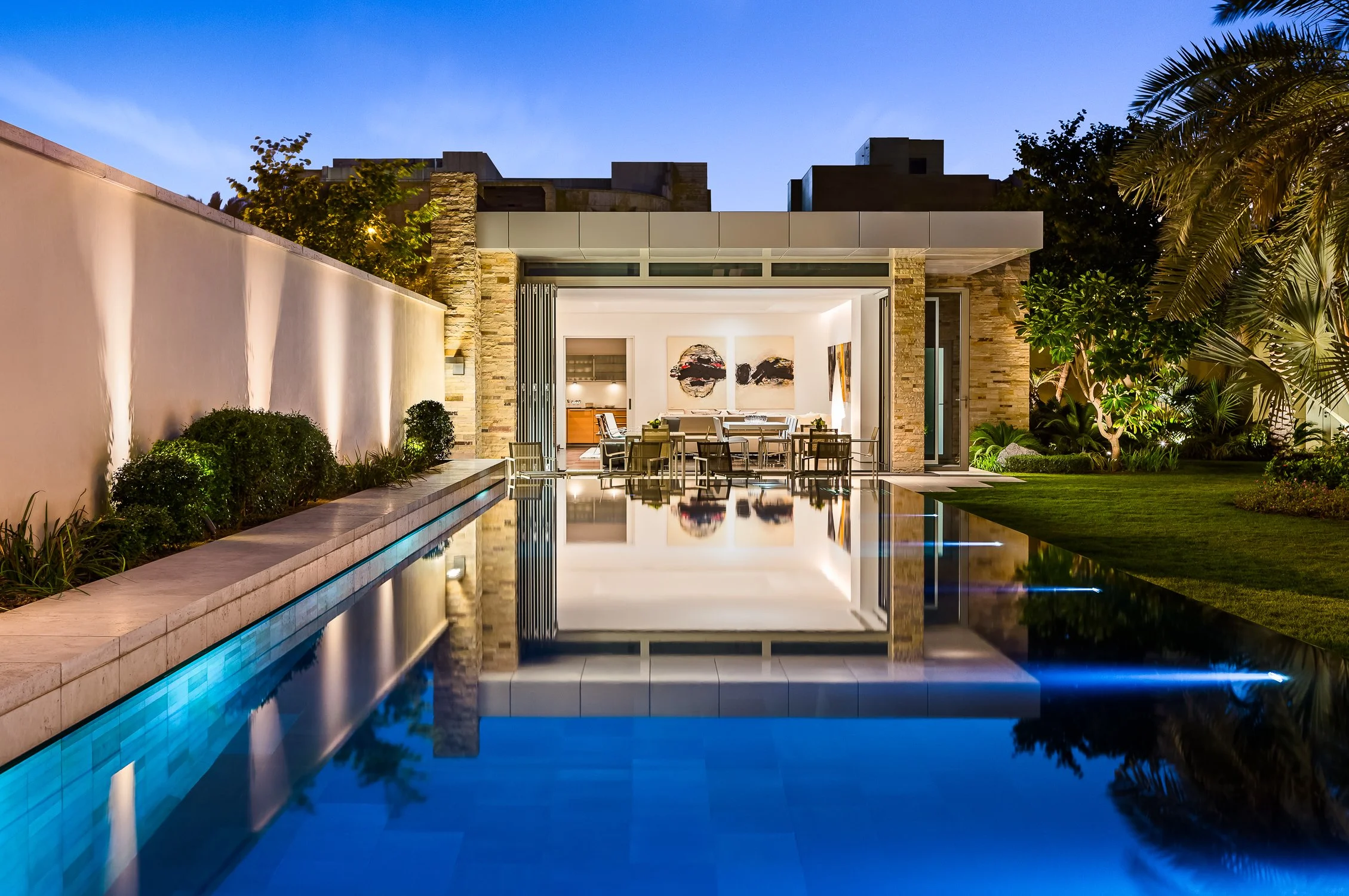 Modern luxury outdoor pool and dining pavilion at Kuwait Estate, featuring sleek architectural design and lush landscaping, illuminated at dusk.