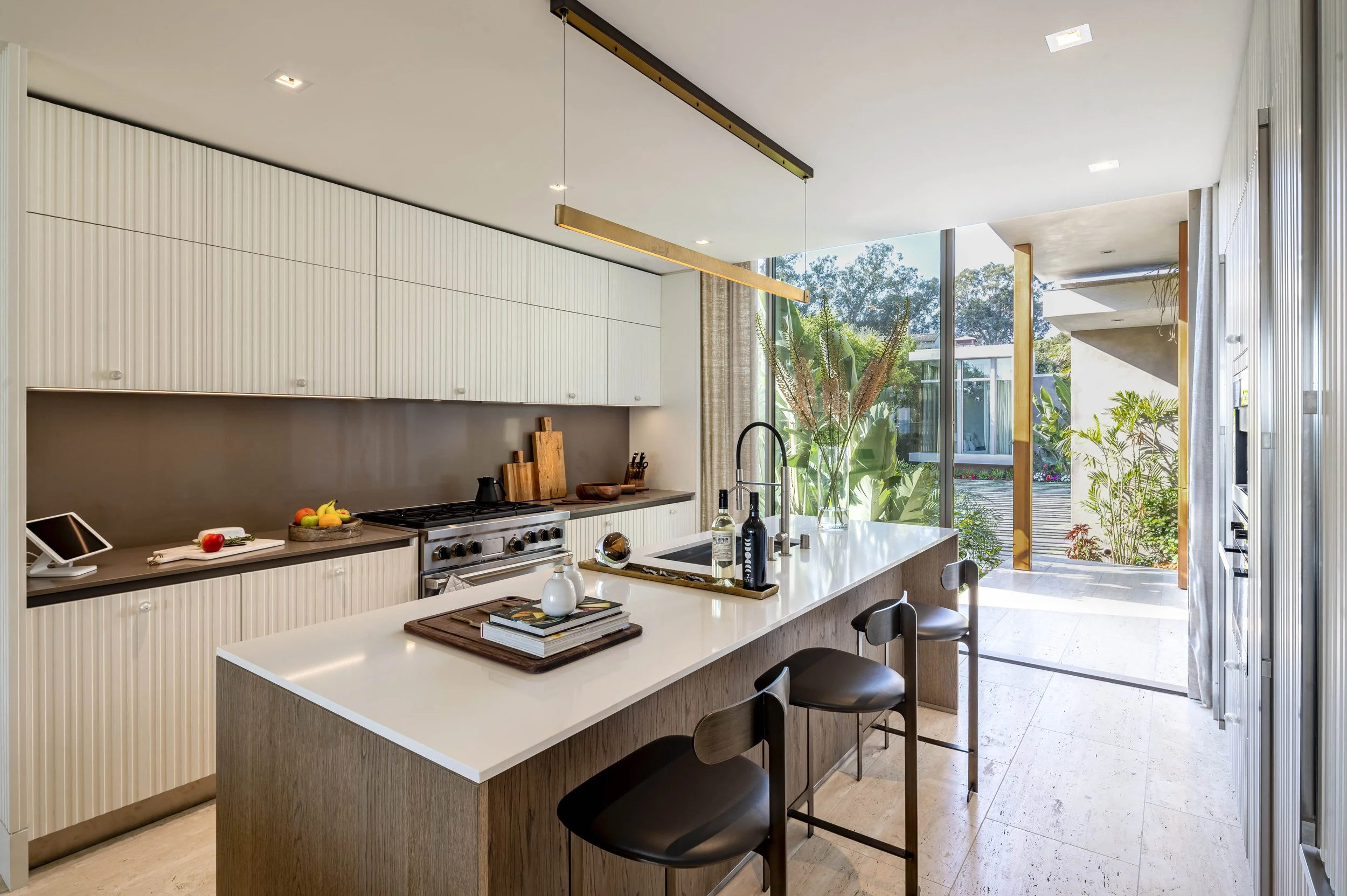 Modern kitchen with large island, bar stools, stainless steel appliances, and glass doors opening to patio with plants.