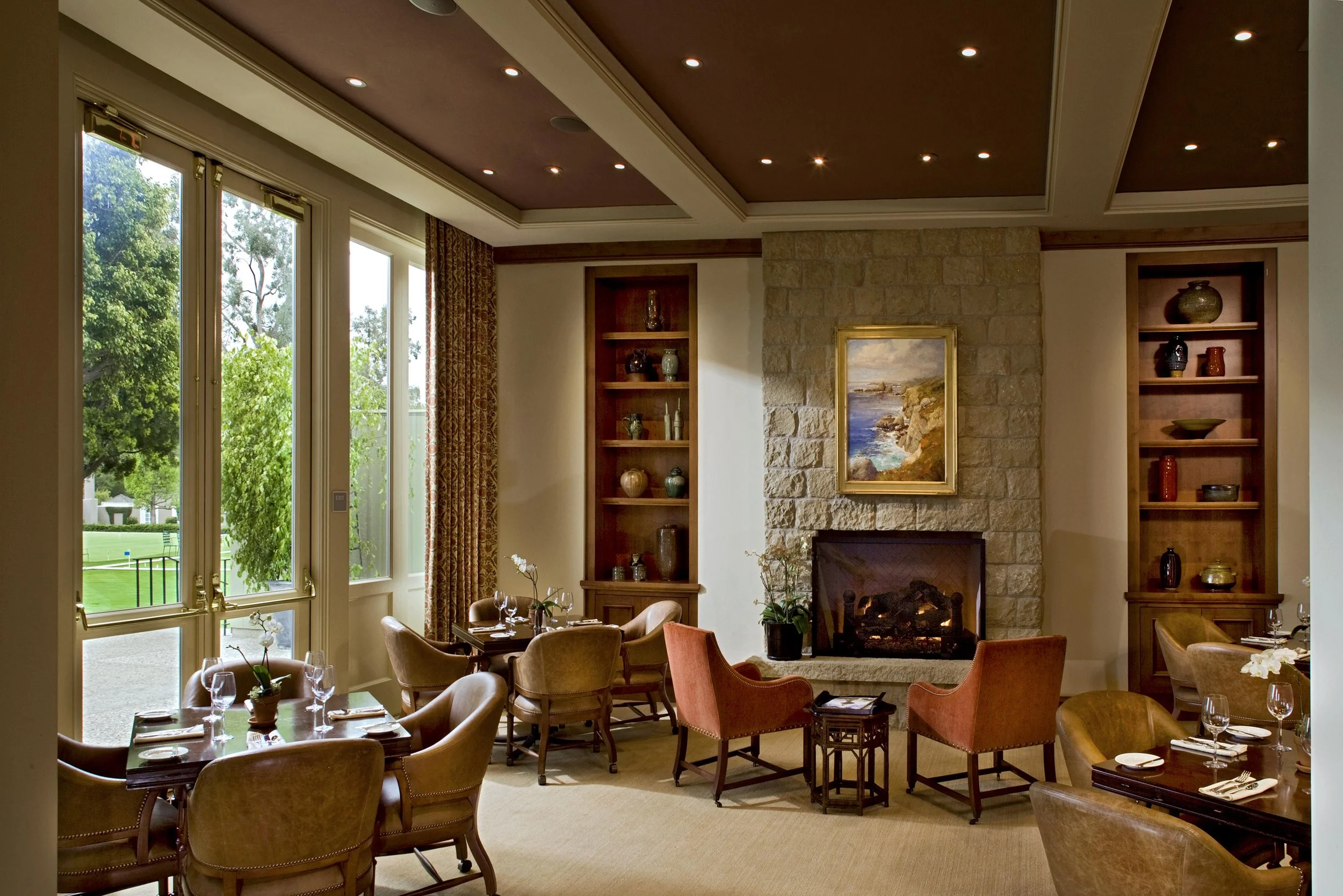 Elegant room with a fireplace, armchairs, dining tables set for a meal, large windows, and decorative shelves.
