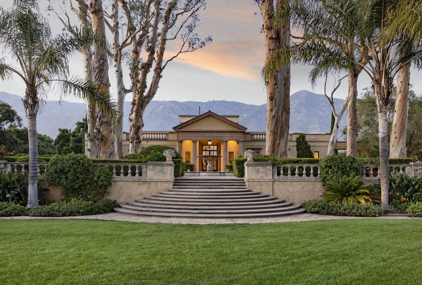 Grand mansion with a landscaped garden and large stone staircase, surrounded by tall trees under a sunset sky.