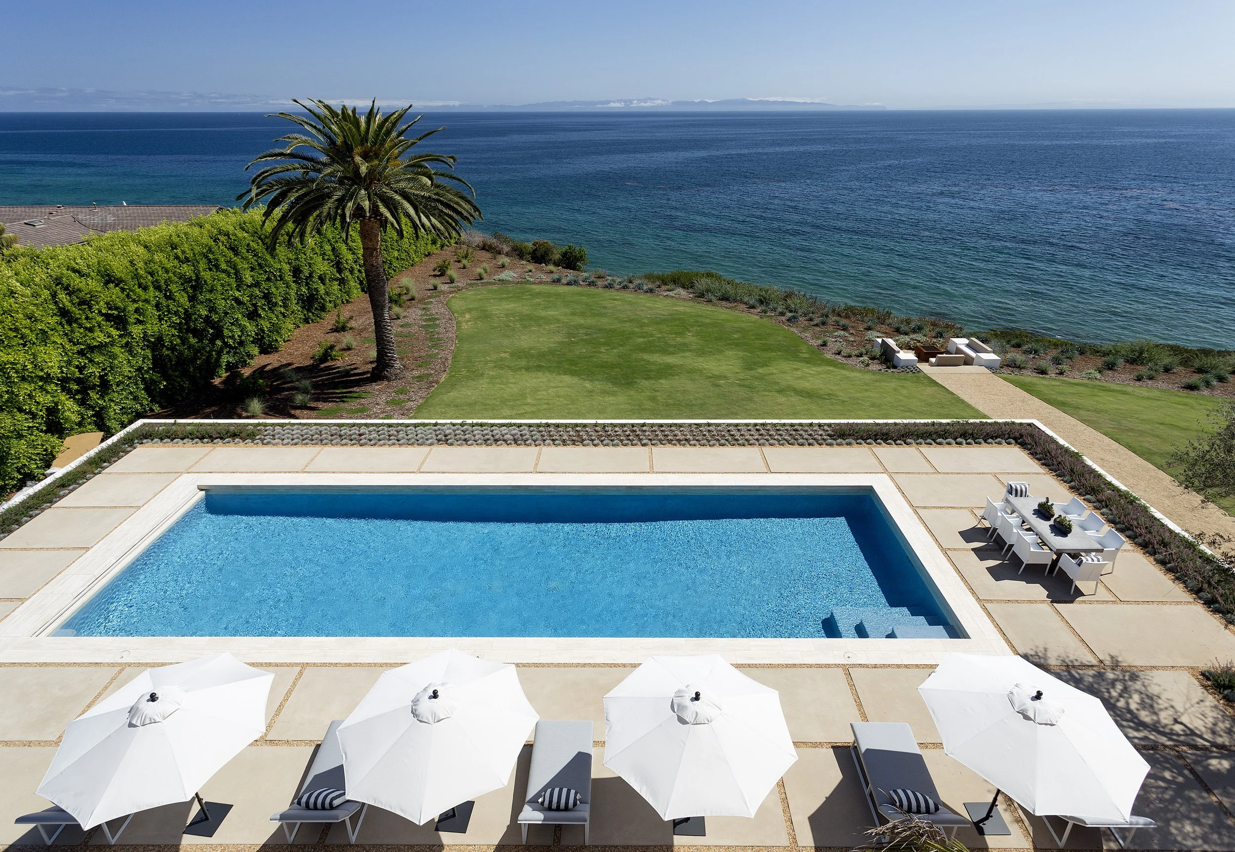 Luxury oceanfront pool with patio, white umbrellas, loungers, dining table, green lawn, palm tree, and ocean view.