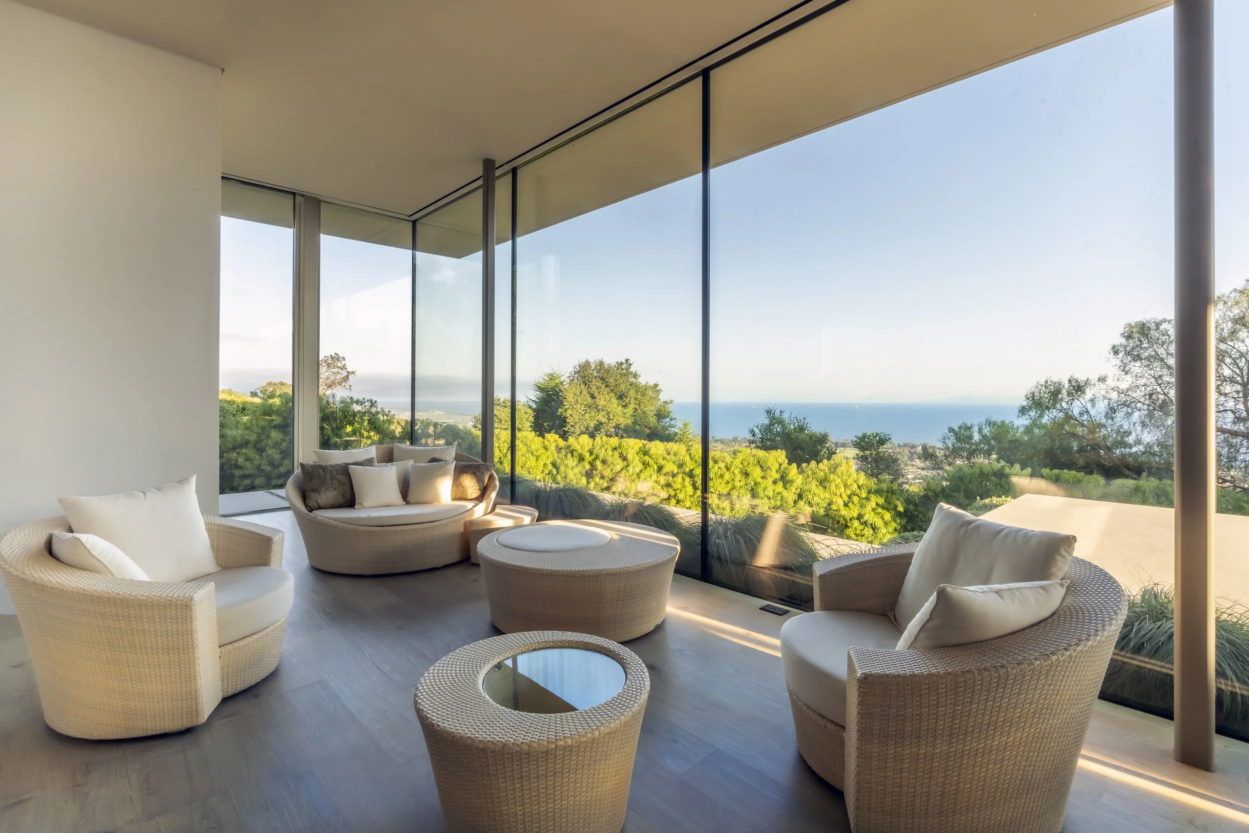 Modern living room with wicker furniture and floor-to-ceiling windows overlooking ocean and greenery.
