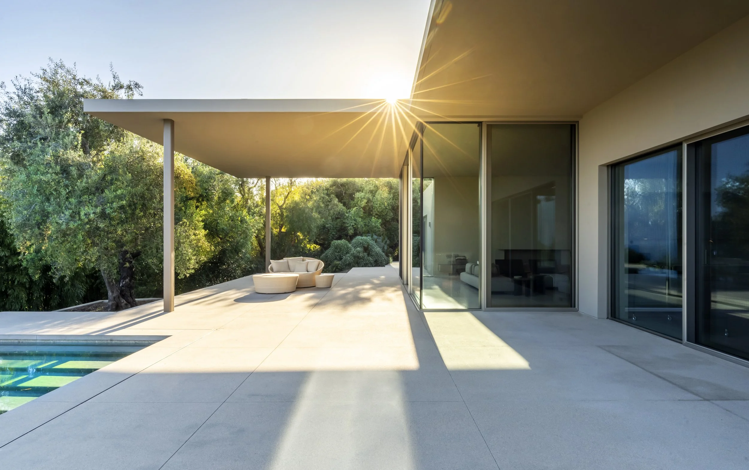 Modern outdoor patio at Toro Canyon Residence by The Warner Group Architects, Inc., featuring minimalist design, comfortable seating, and large glass windows with sunlight streaming through the trees.