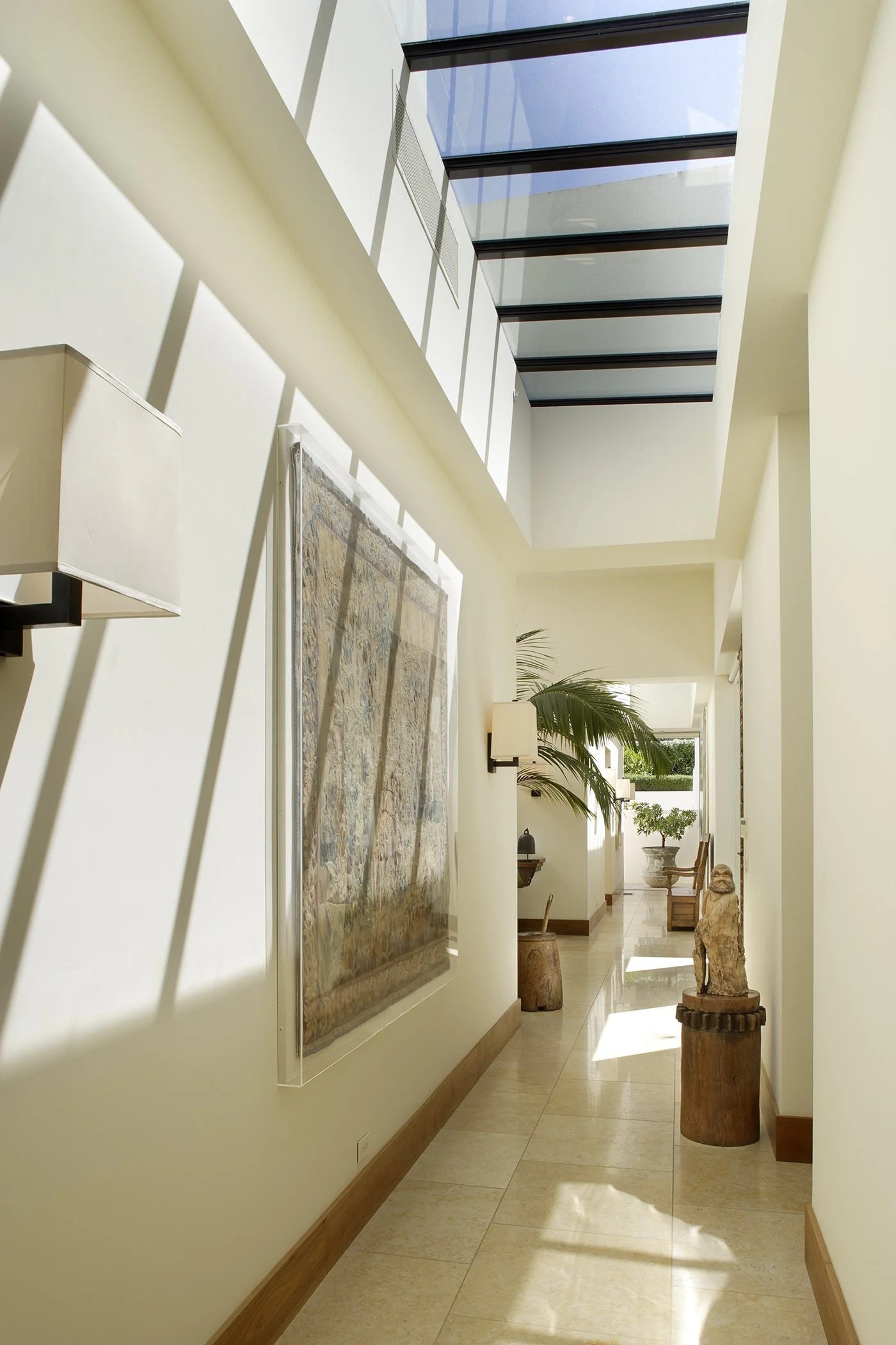 Modern hallway with glass ceiling, wall art, sconces, plant, and sculptures.