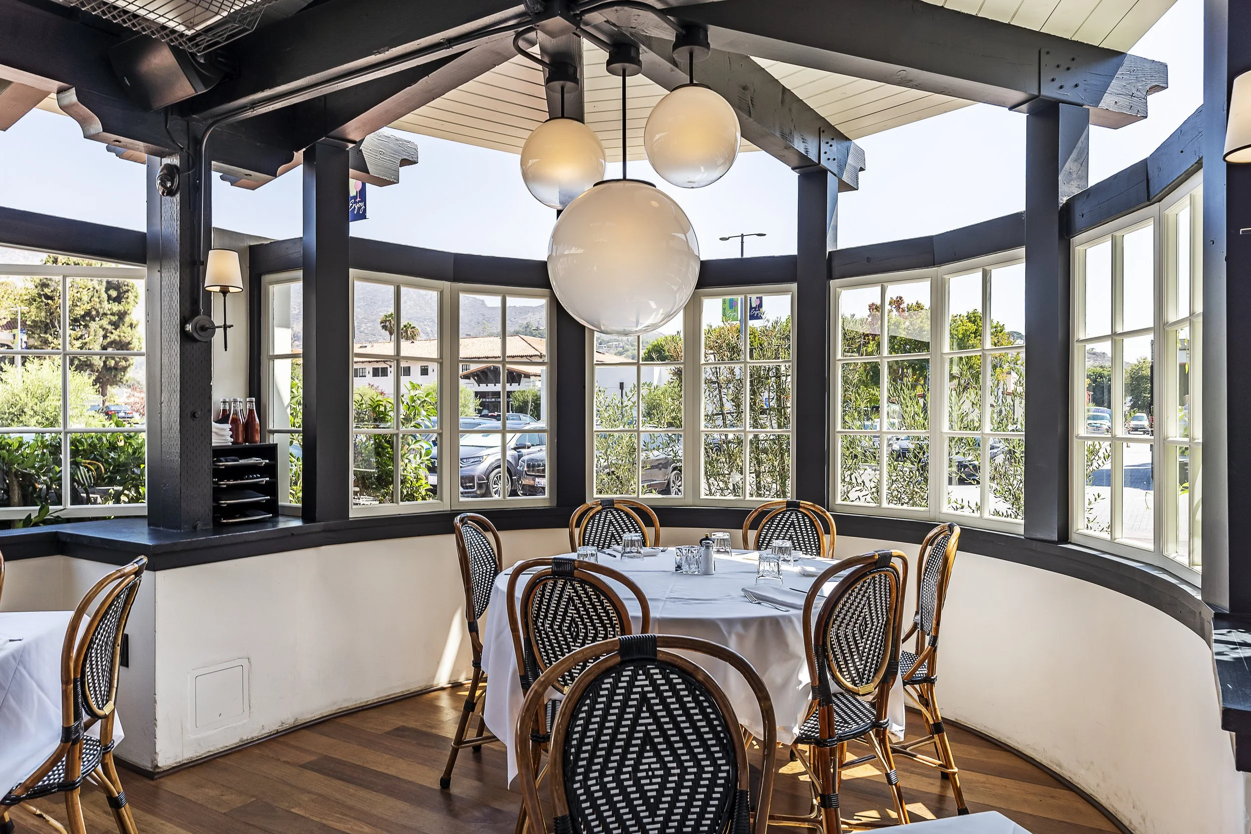 Elegant dining area with large windows, round table, and modern lighting at Lucky’s Steakhouse in Malibu, offering views of the Malibu Country Mart.