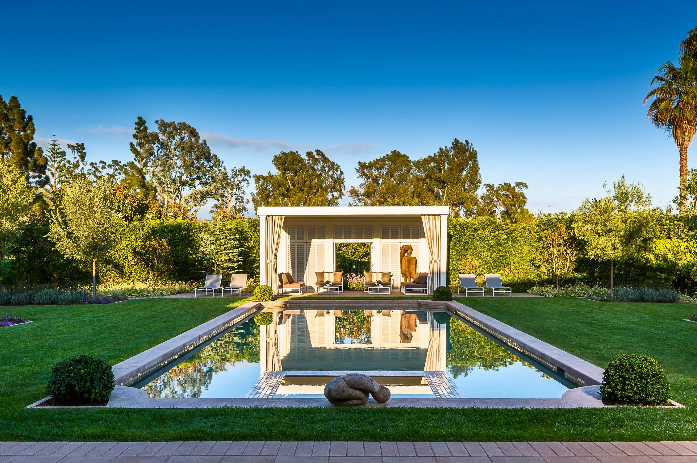 Elegant backyard with a rectangular pool, surrounded by manicured grass and shrubs, facing a modern cabana with patio furniture, trees in the background.