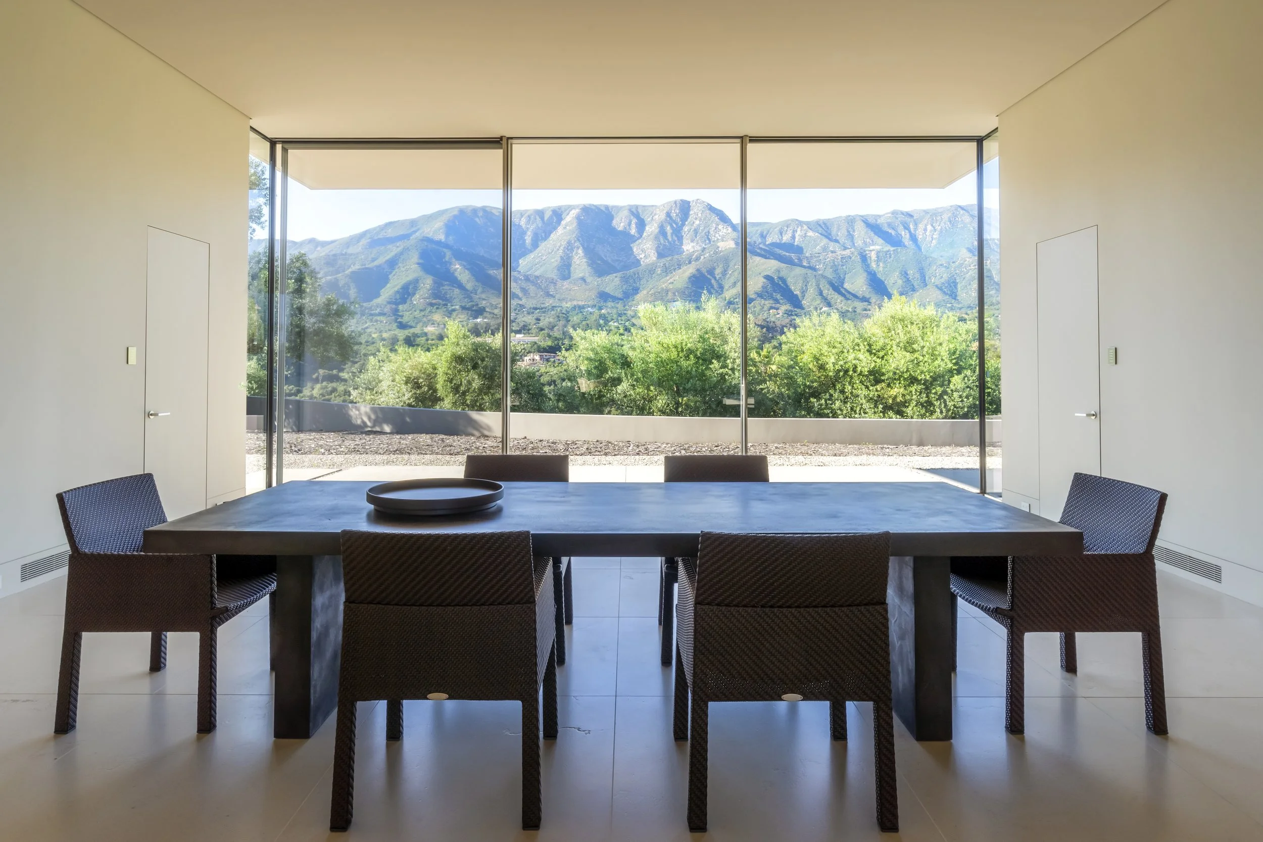 Dining area at Toro Canyon Residence by The Warner Group Architects, Inc., featuring a minimalist design with floor-to-ceiling windows offering panoramic views of the mountains and surrounding landscape.
