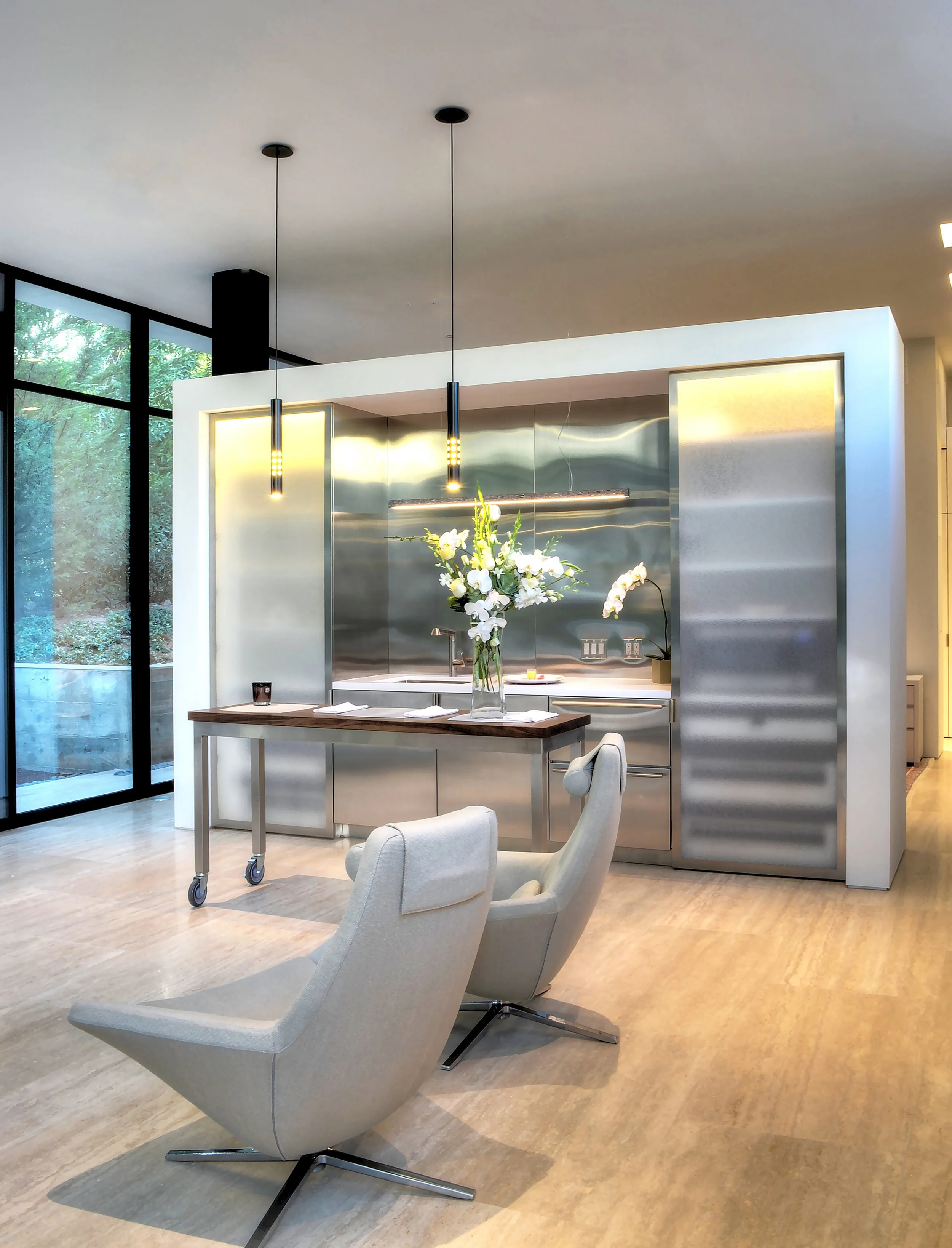 Modern kitchen with stainless steel appliances, minimalist island table, and lounge chairs, featuring large glass windows overlooking greenery, designed by The Warner Group Architects, Inc.