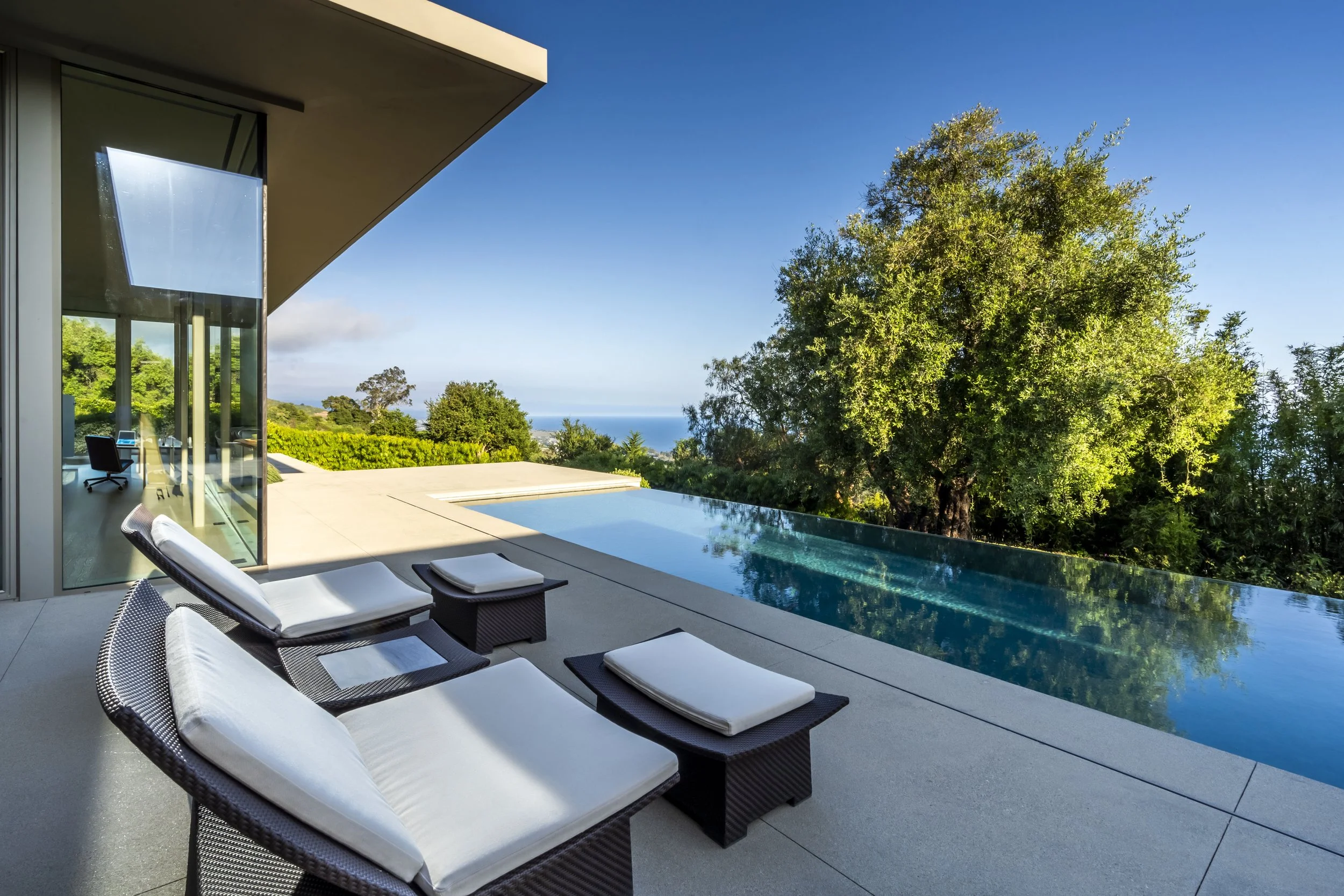 Modern outdoor patio with lounge chairs next to an infinity pool, surrounded by trees and ocean view in the background.