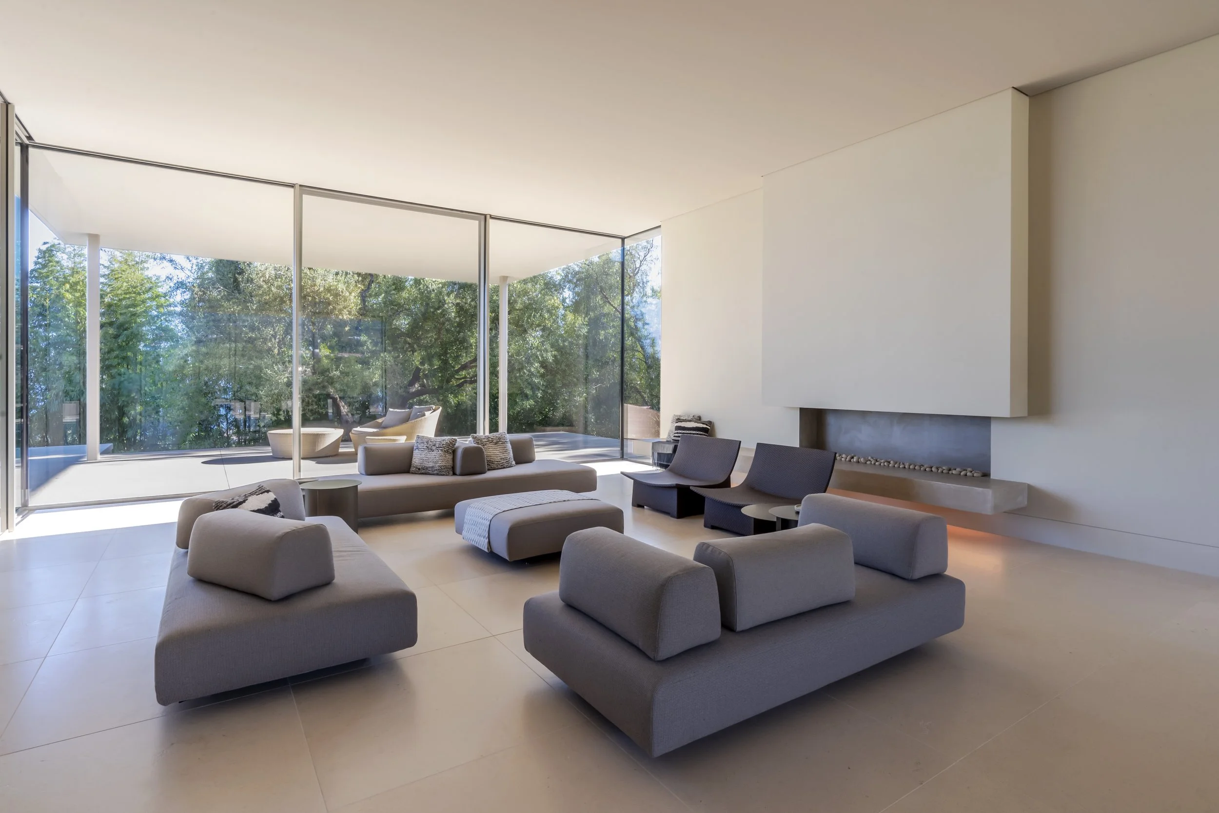 Modern living room with minimalist furniture, large windows, and a sleek fireplace. The space opens to an outdoor area with trees visible.
