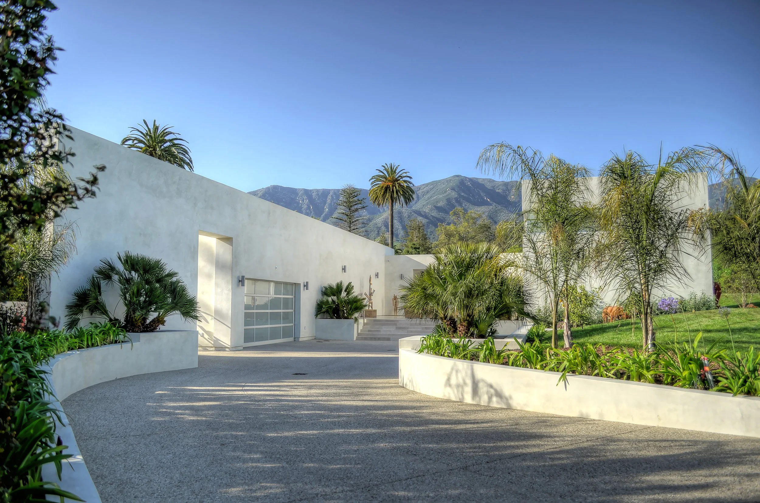 Modern white building with palm trees, landscaped garden, and mountain backdrop under clear blue sky.