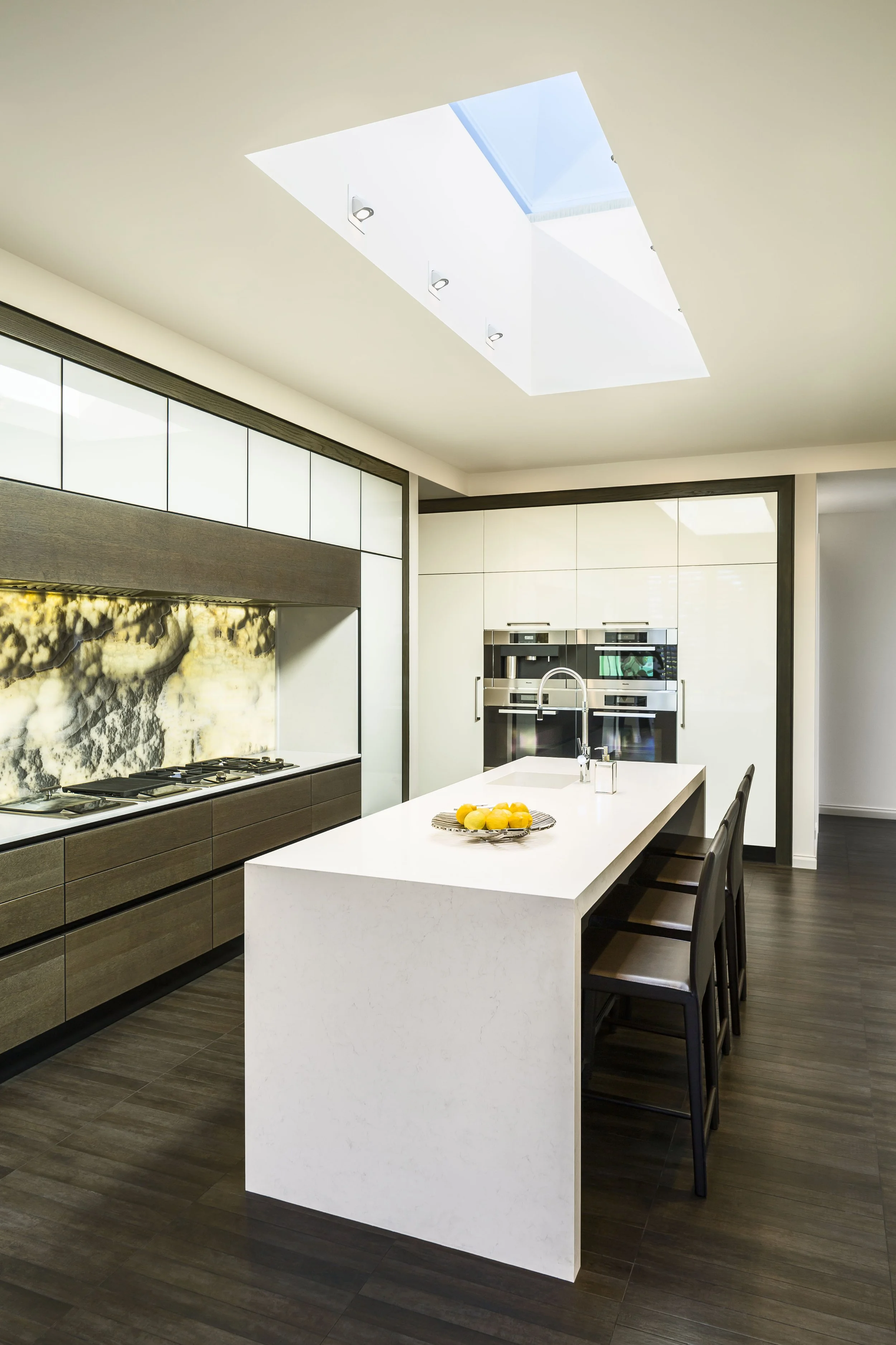 Modern kitchen with skylight, featuring sleek white countertops, a marble backsplash, integrated cabinetry, and a central island with a stainless steel sink at the Birnam Wood Residence.