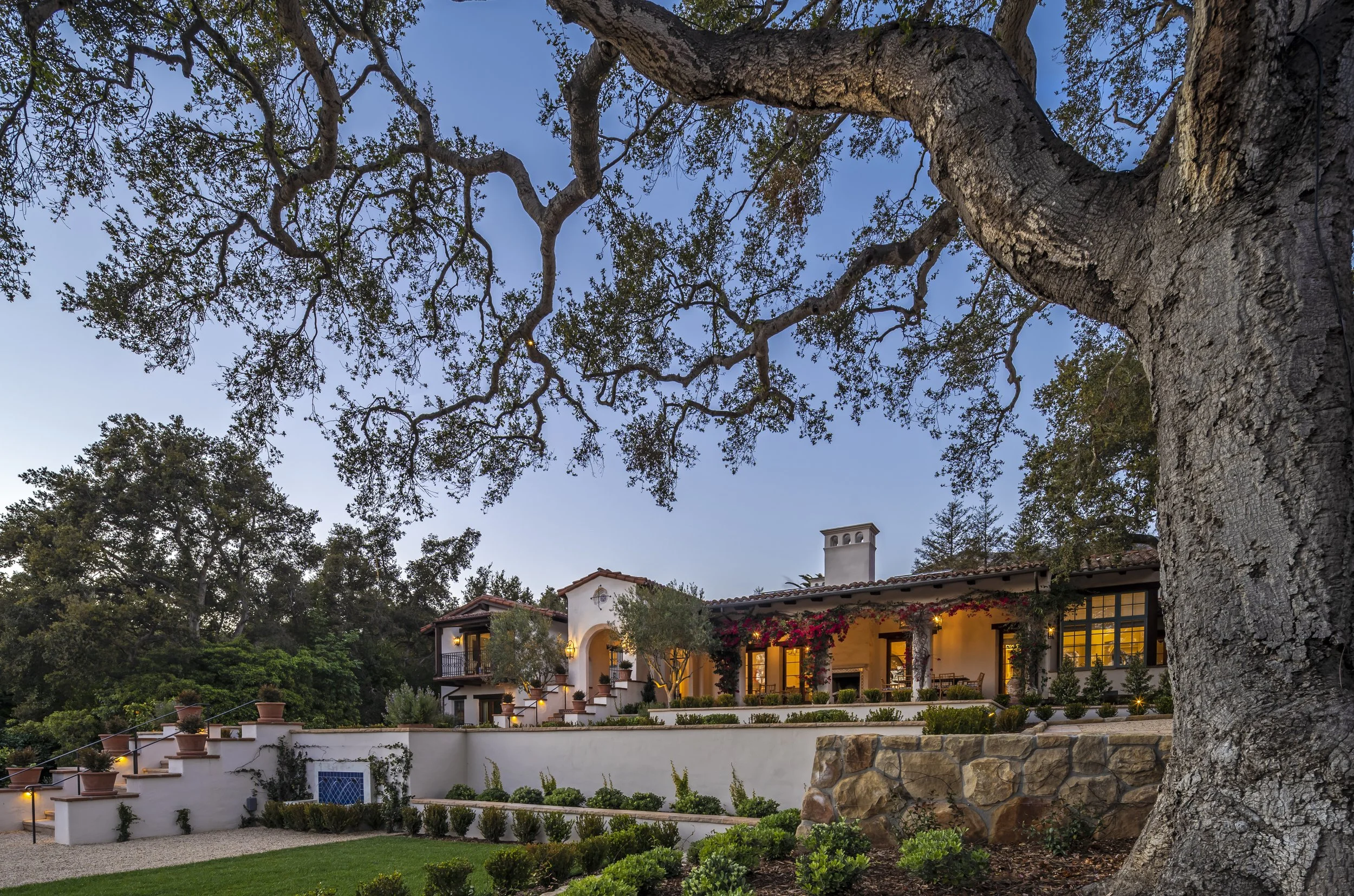 Elegant Mediterranean-style villa surrounded by lush greenery, with a large tree in the foreground under a twilight sky.