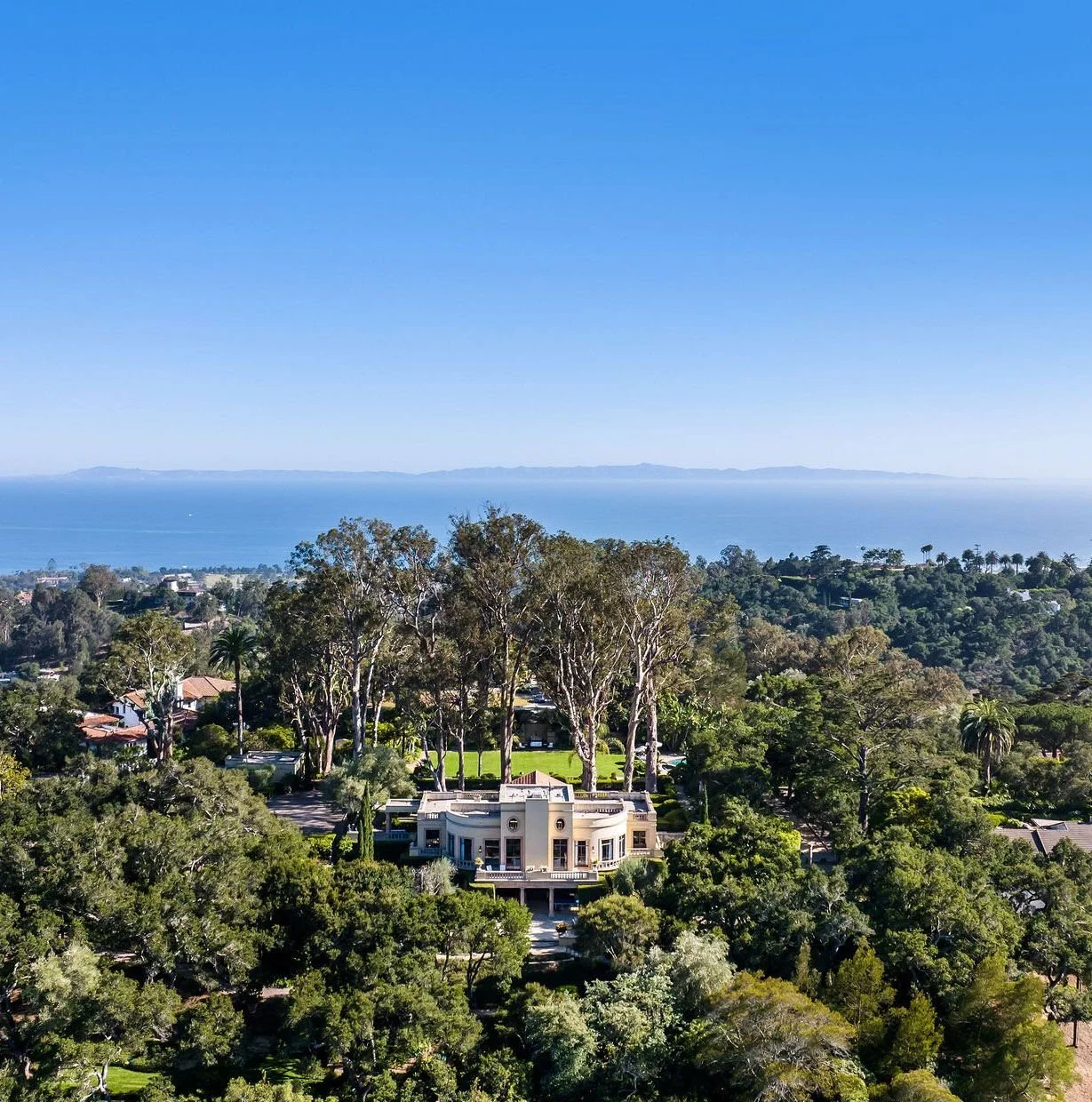 Aerial view of a large house surrounded by trees with an ocean and distant mountains in the background.