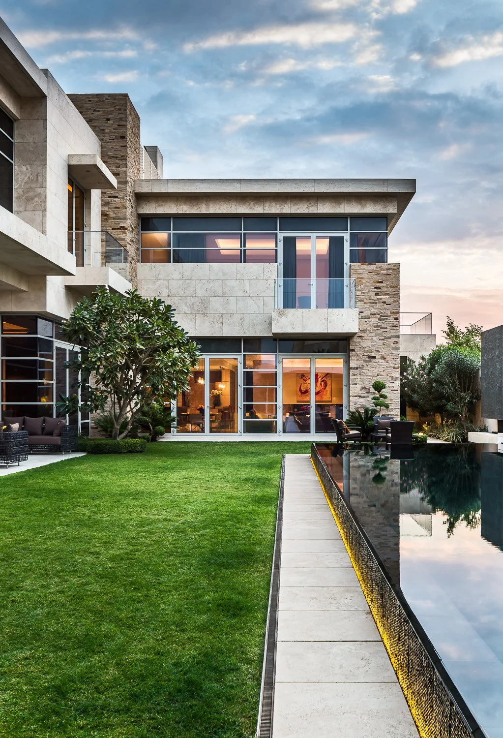 Modern stone exterior of a luxury estate in Kuwait with large glass windows, lush greenery, and a sleek infinity pool, blending contemporary architecture with natural surroundings.