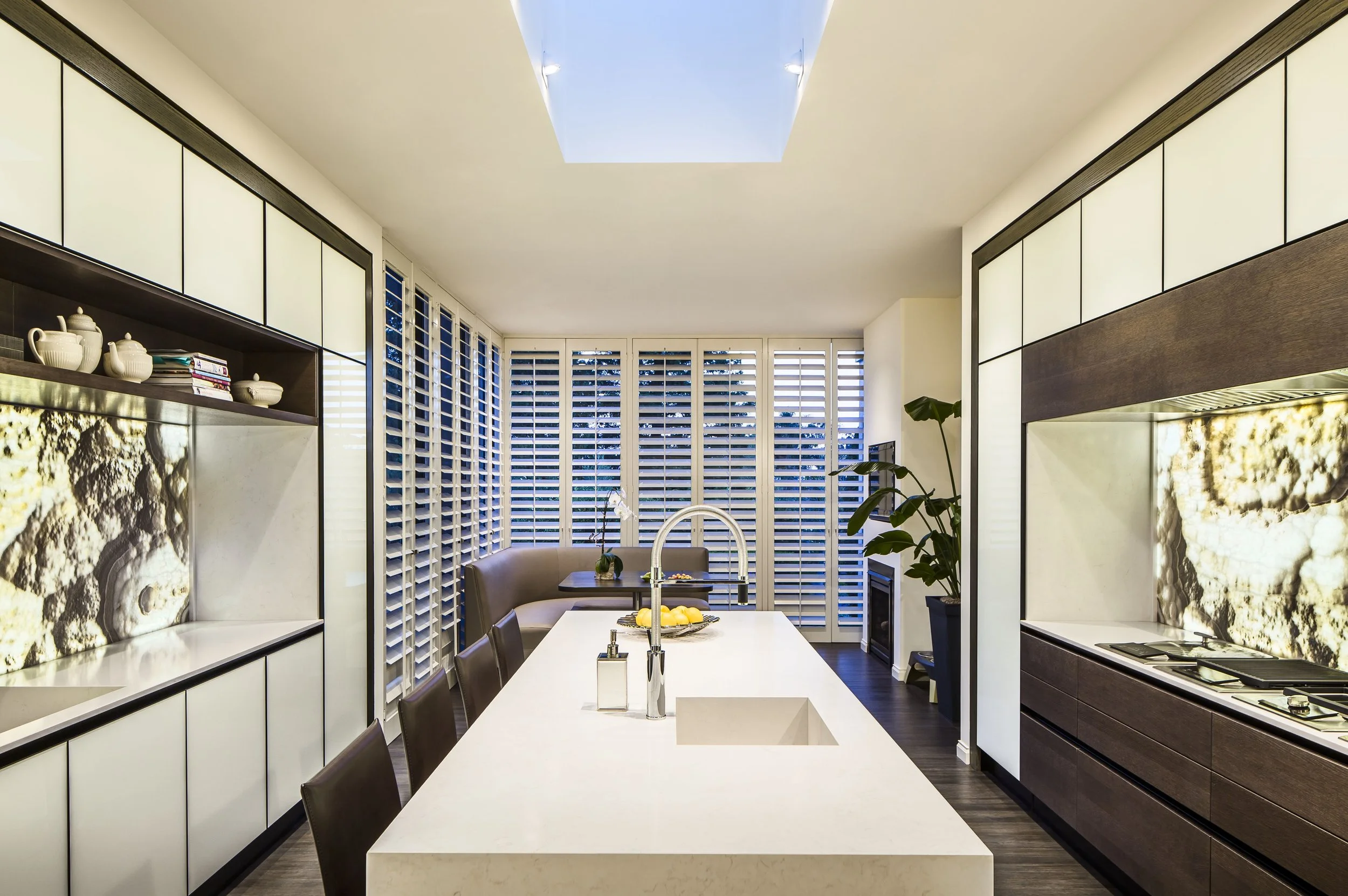 Modern kitchen with a long white island, dark chairs, built-in appliances, marble backsplash, indoor plant, and large windows with white shutters.