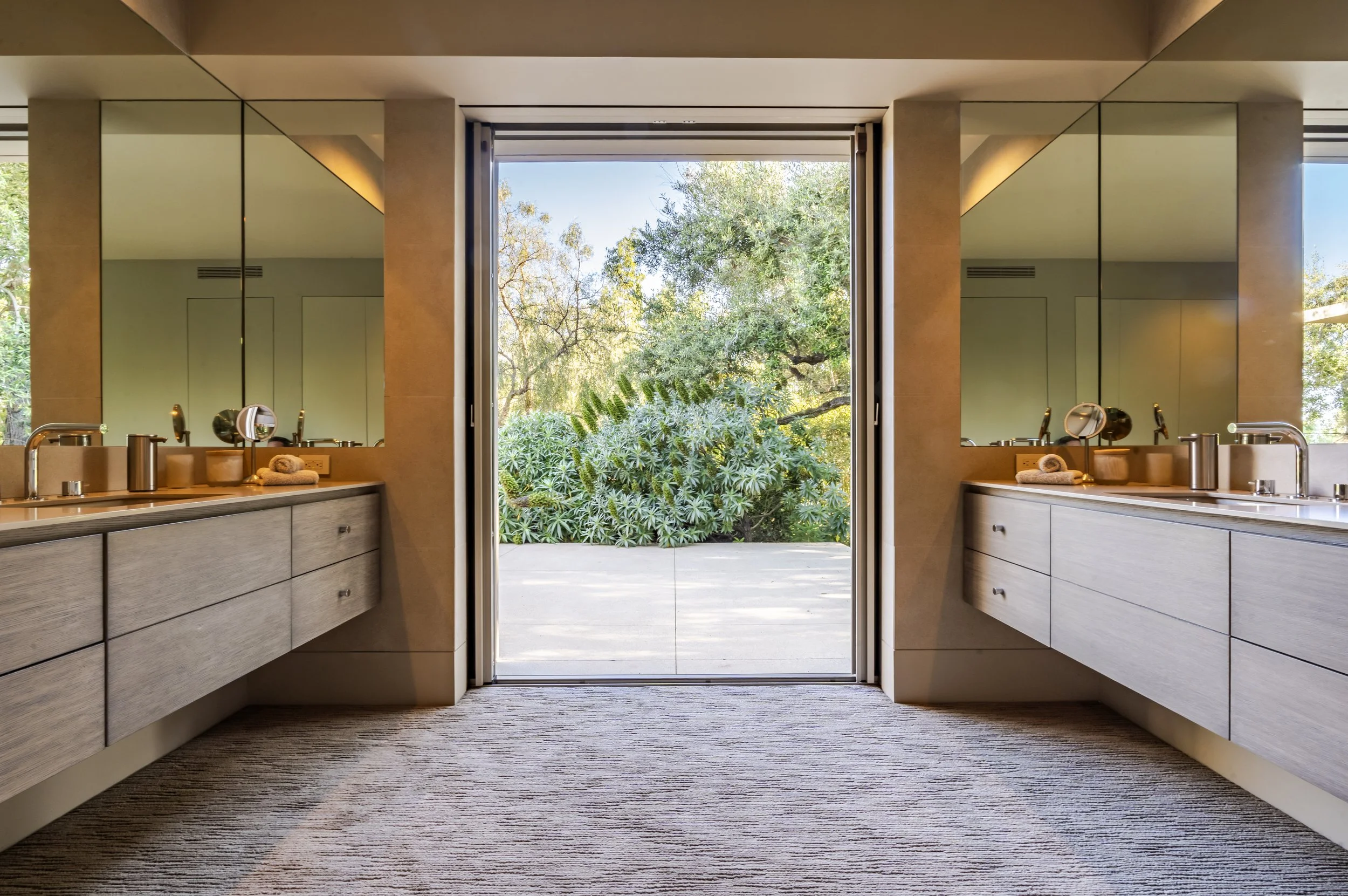 Luxury bathroom at Toro Canyon Residence by The Warner Group Architects, Inc., featuring dual vanities with modern mirrors and a large sliding door opening to lush outdoor views, blending indoor and outdoor living.