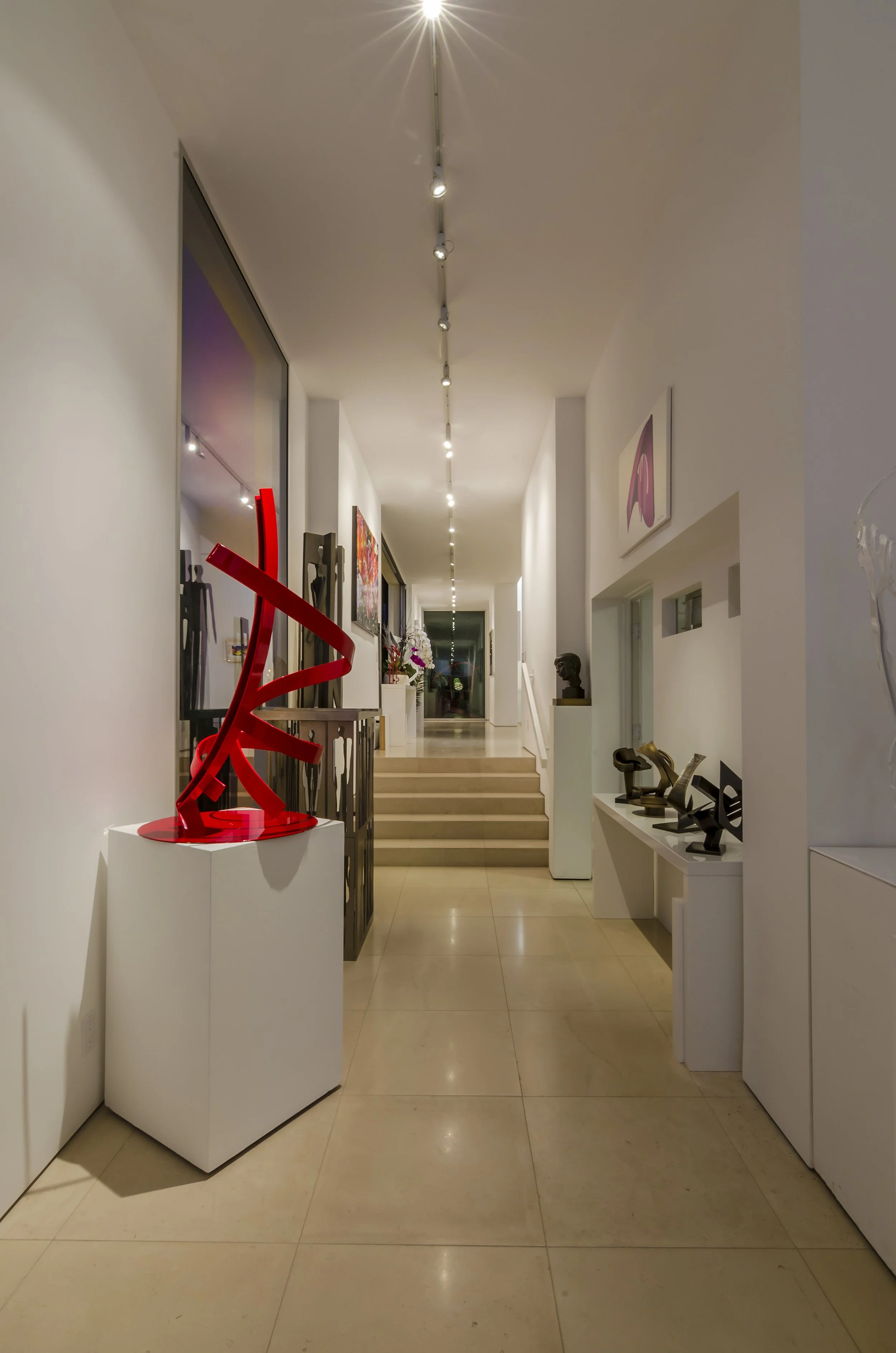 Art gallery hallway with modern sculptures and paintings on display, featuring a bright red abstract sculpture on a pedestal.