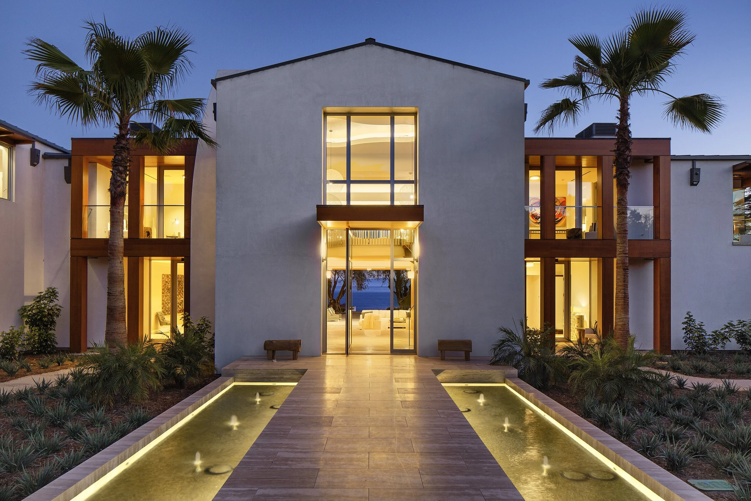 Modern architectural facade with glass accents and symmetrical lines, featuring a grand entryway, flanked by palm trees and reflecting pools, offering a seamless blend of luxury and nature.