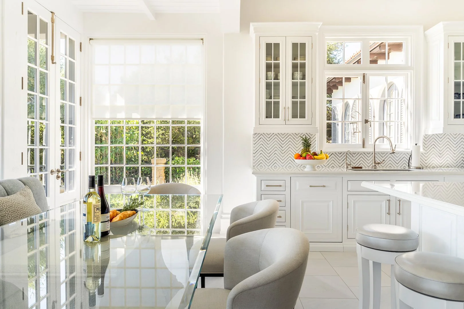 Modern white kitchen with large windows, glass table, cushioned chairs, fruit bowl, and wine bottles.