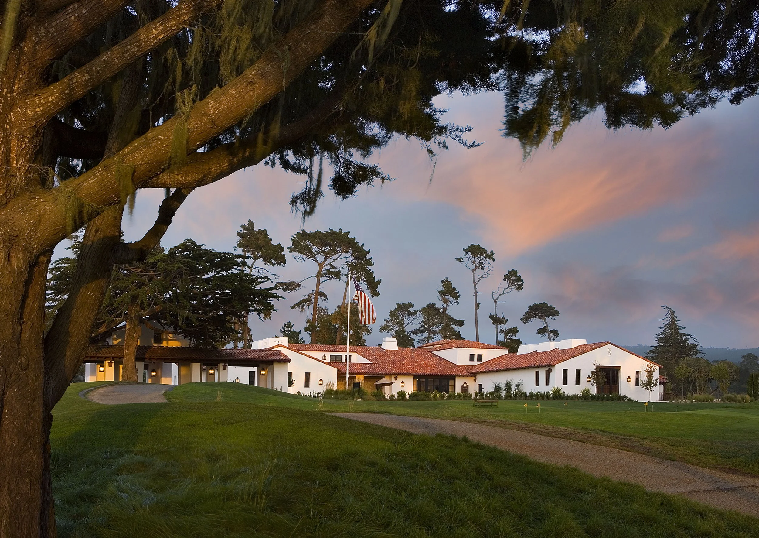A Spanish-style villa with a red tile roof and white walls, surrounded by trees and a lush lawn. An American flag is displayed in front of the house, under a colorful sunset sky.