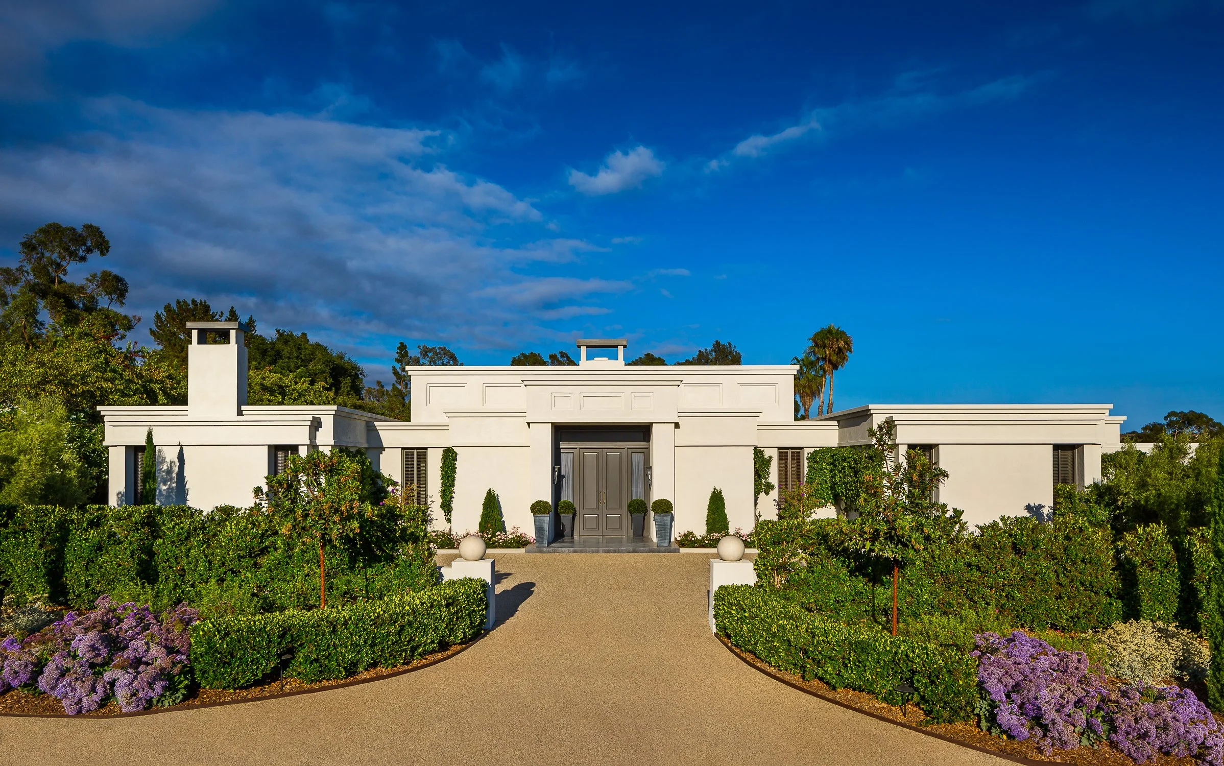 Modern Mediterranean-style white luxury home surrounded by lush greenery, showcasing a grand entrance with symmetrical landscaping and clean architectural lines, designed by The Warner Group Architects, Inc. at Birnam Wood Residence.