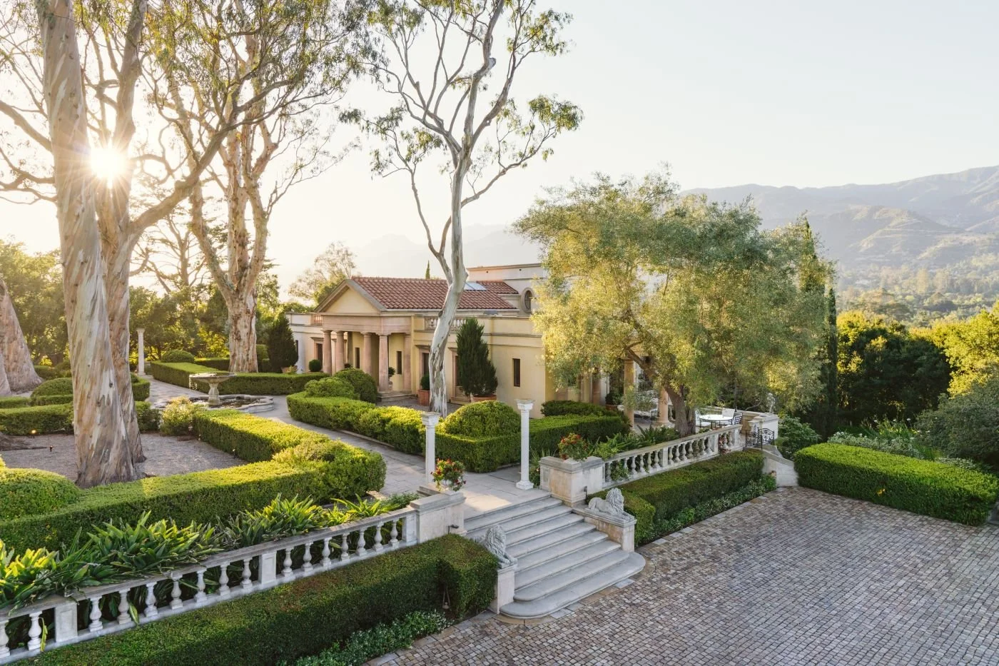 Elegant estate with manicured gardens, grand staircase, and classical architecture surrounded by lush greenery and mature trees, mountains in background.