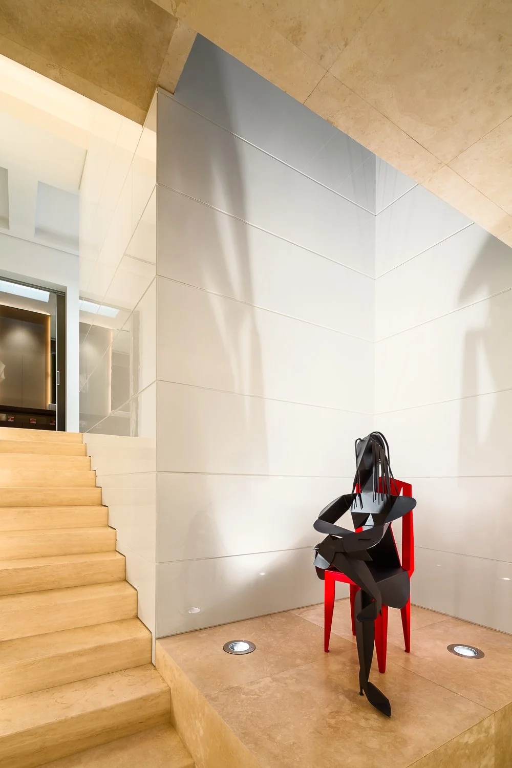 Modern art installation featuring an abstract black sculpture seated on a vibrant red chair, positioned at the base of a light stone staircase in a sleek, minimalistic interior.