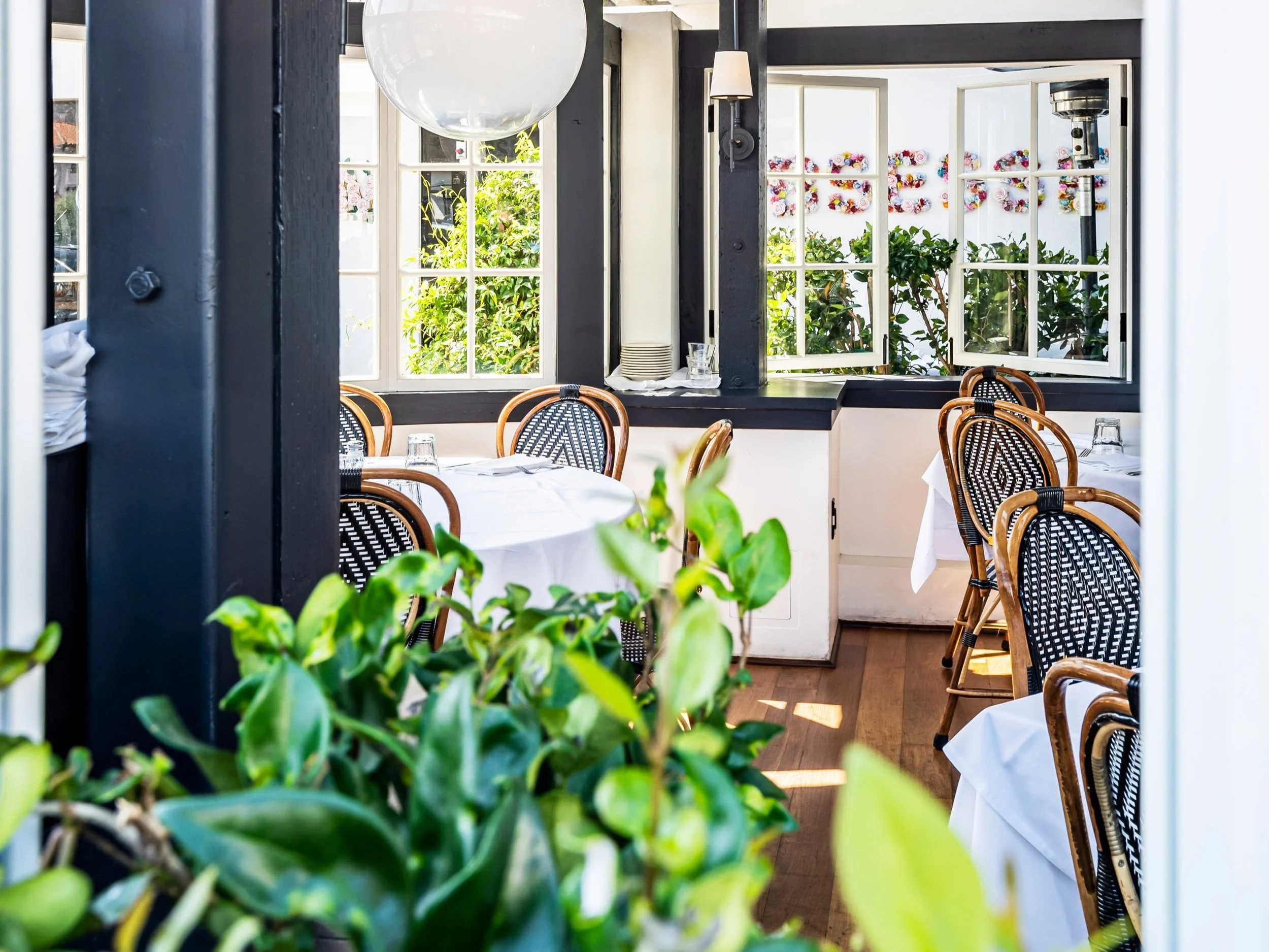 Bright and airy restaurant interior with wicker chairs and white tablecloths, surrounded by windows and greenery.