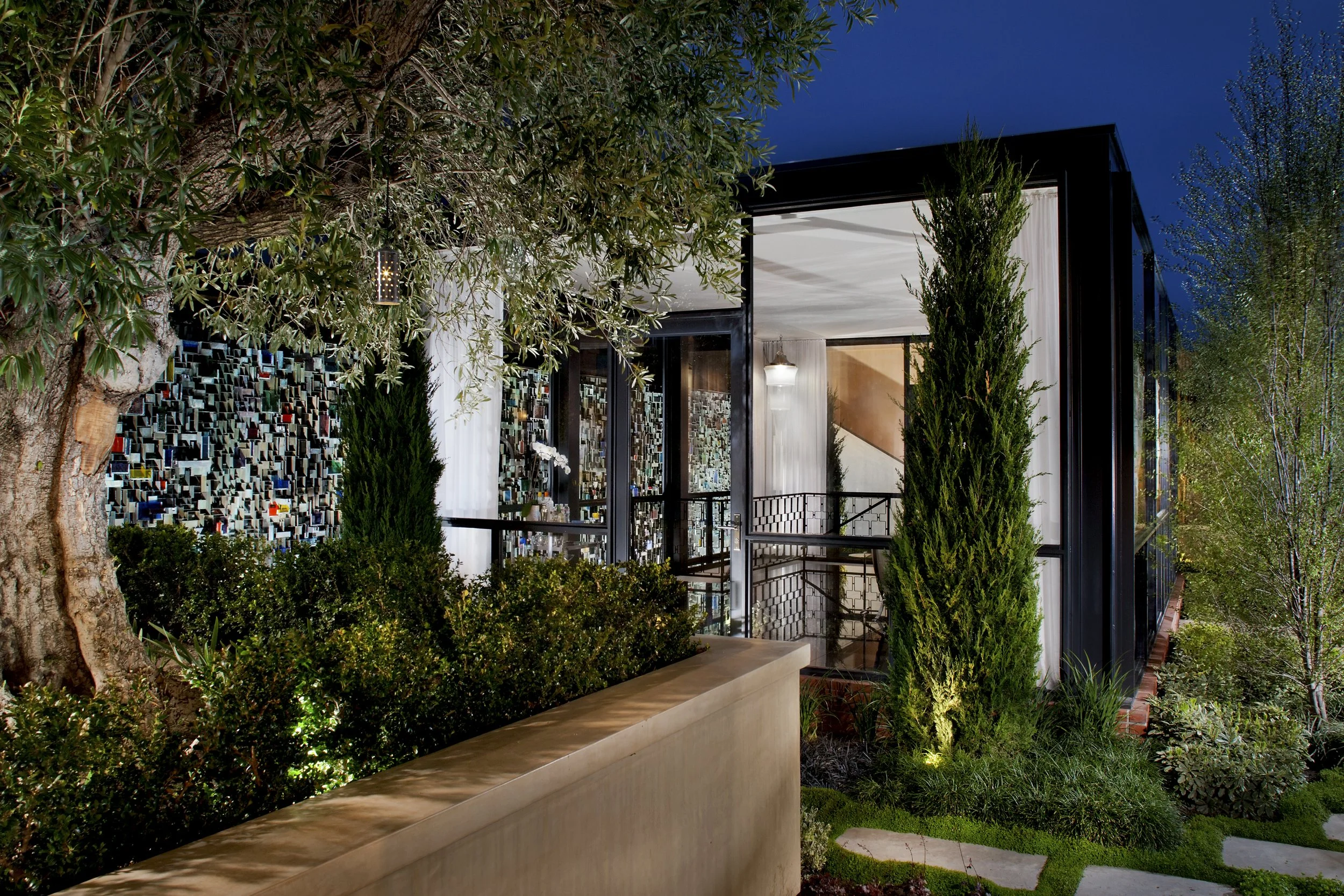 Modern glass house surrounded by trees and greenery at night.