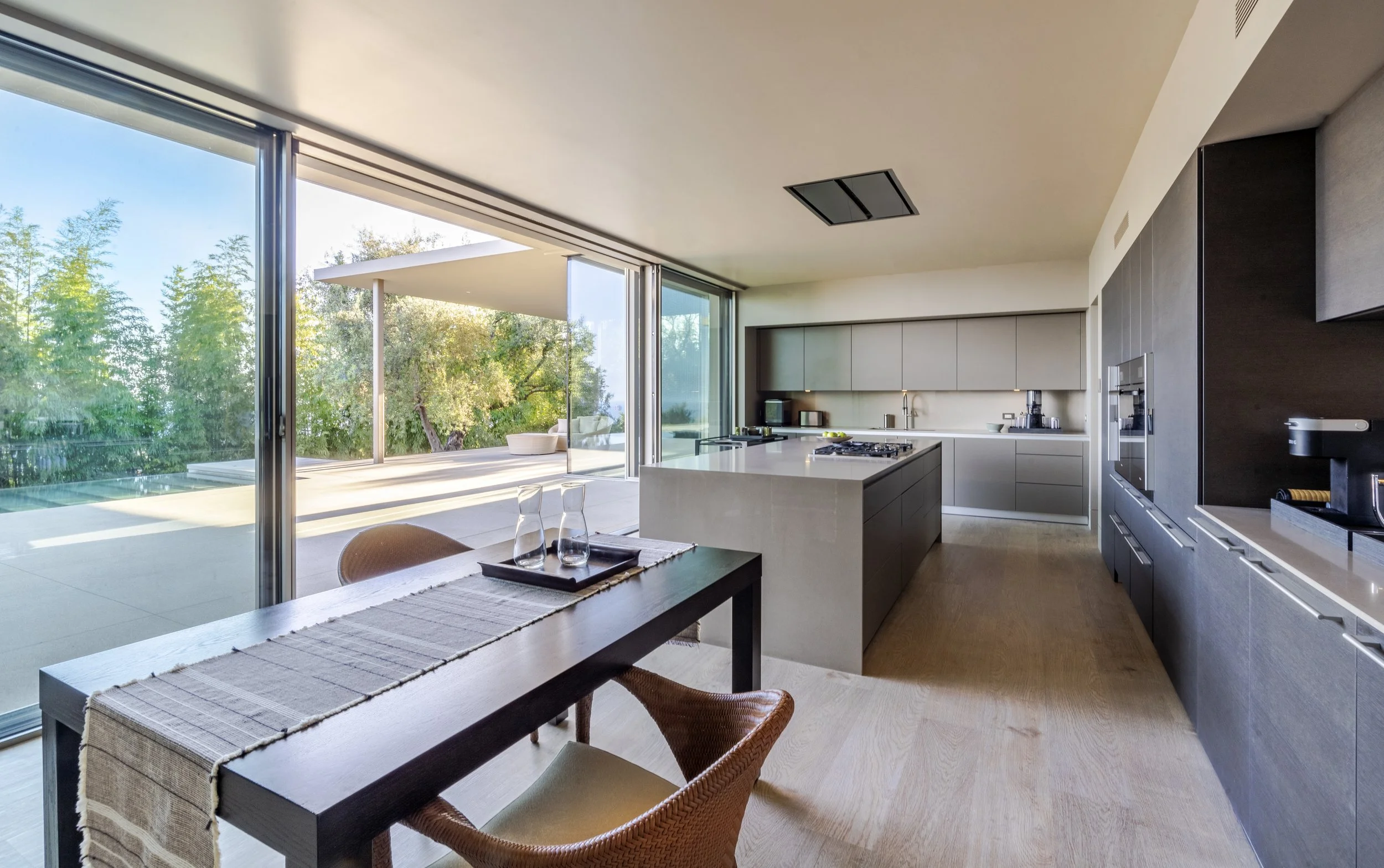 Open-plan kitchen and dining area at Toro Canyon Residence by The Warner Group Architects, Inc., featuring sleek modern cabinetry, large kitchen island, and sliding glass doors that open to an expansive outdoor patio with pool views.