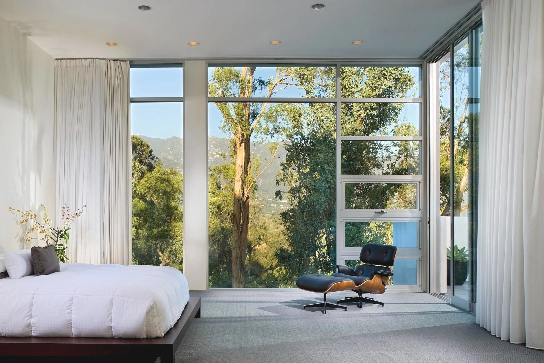 Modern bedroom with large windows, white bedspread, and an Eames lounge chair overlooking trees and mountains.