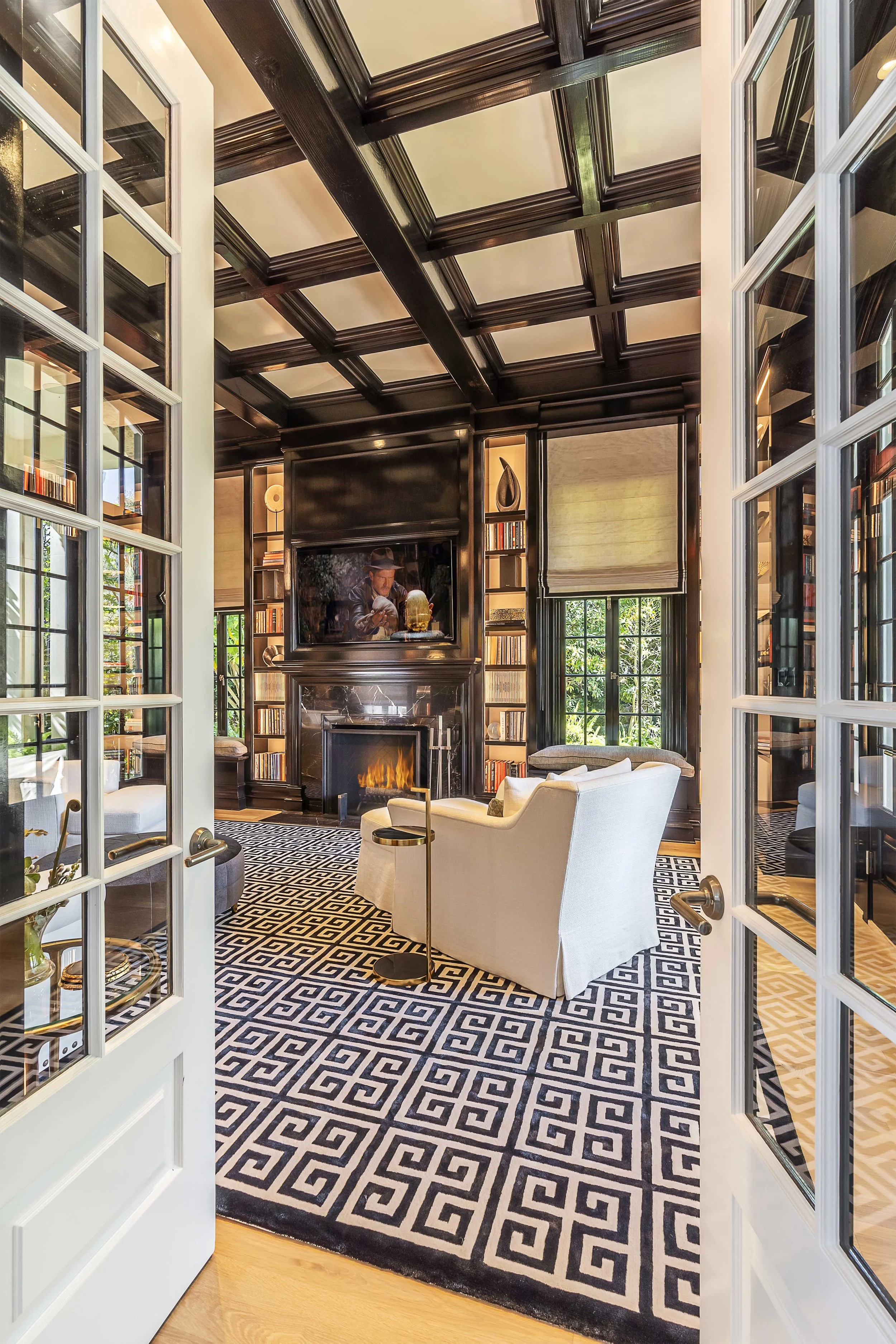 Luxurious home library with coffered ceiling, fireplace, and custom-built shelving, featuring a geometric-patterned rug, designed by The Warner Group Architects, Inc.