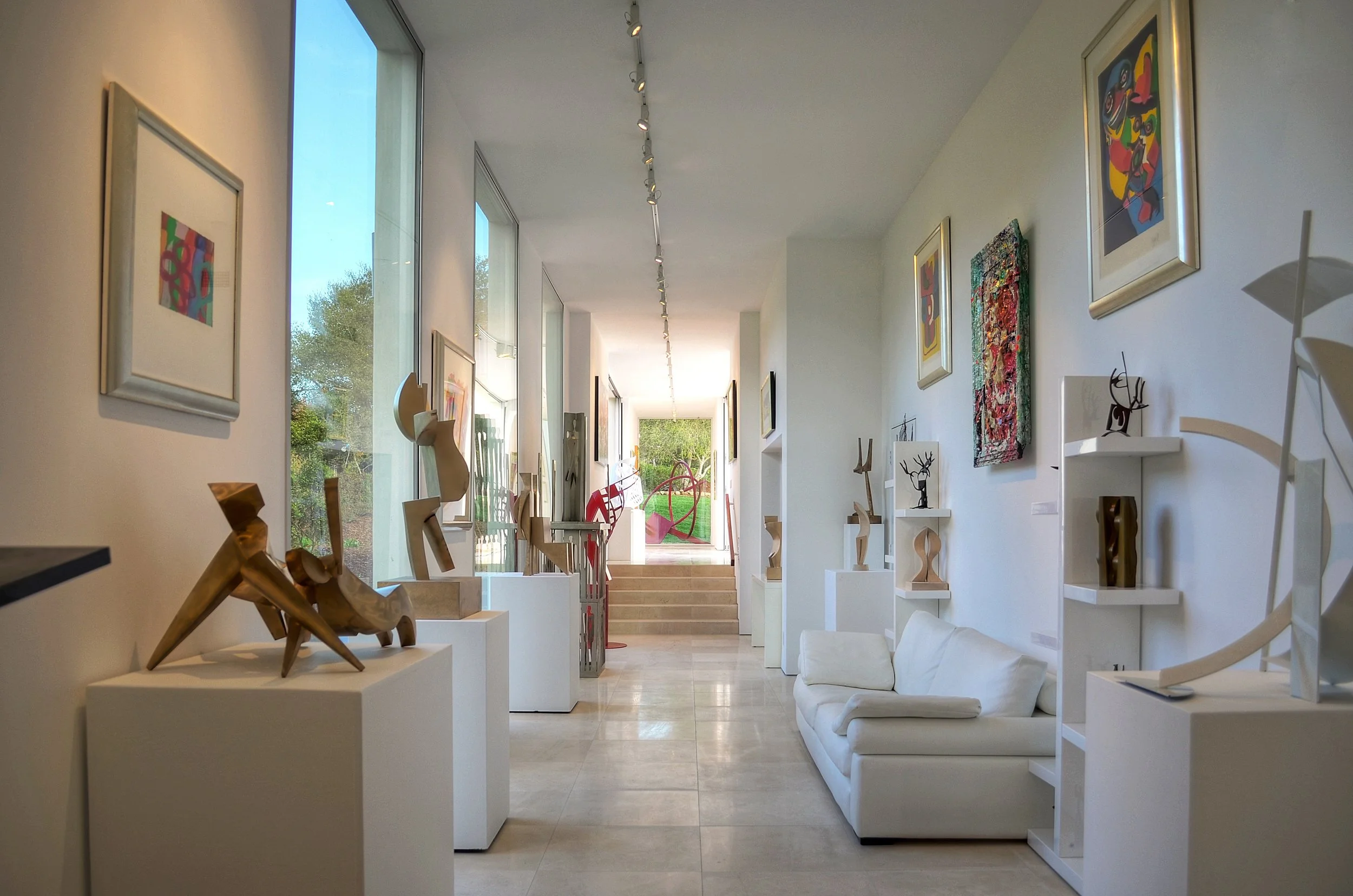Modern art gallery with sculptures and paintings on display, featuring large windows and a white sofa.