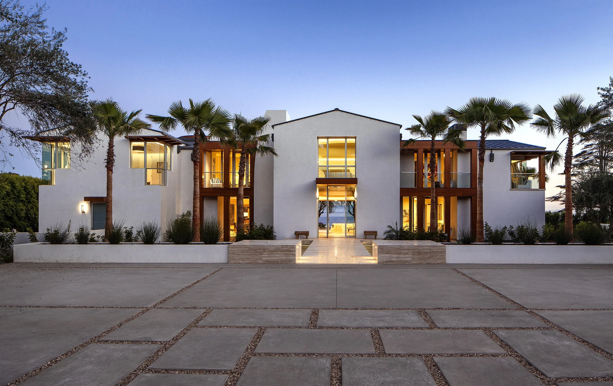 Modern white mansion with palm trees, large windows, and geometric driveway at dusk.
