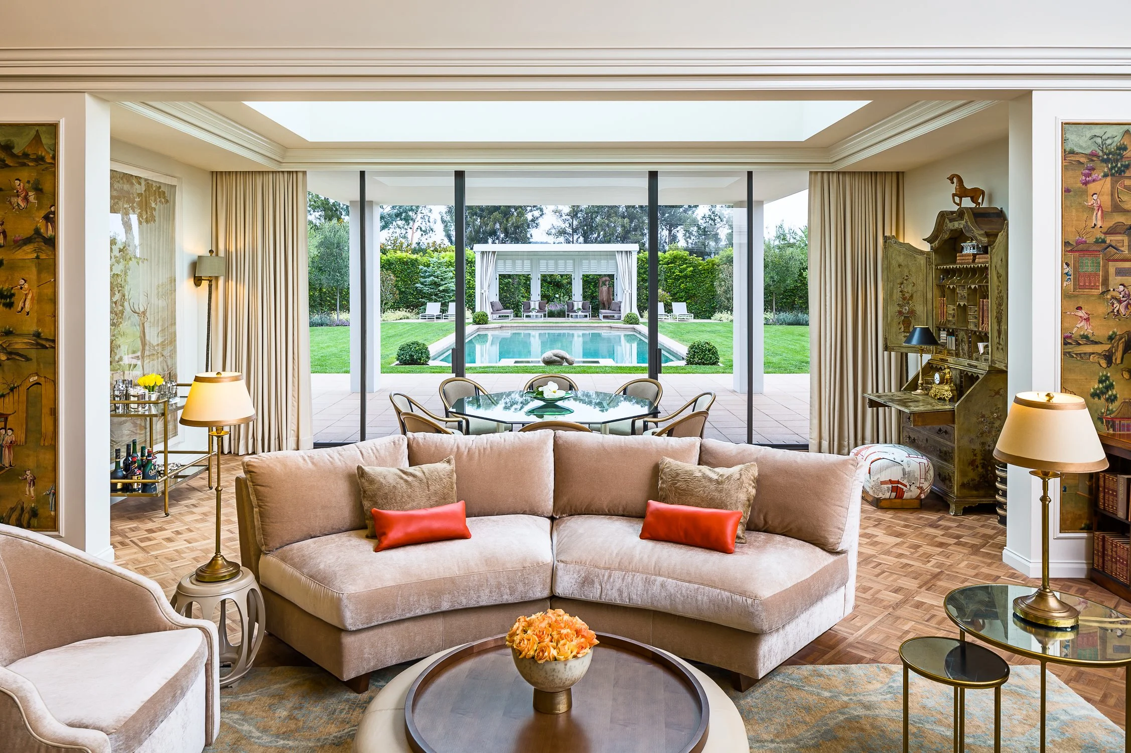 Elegant living room with a large curved couch facing floor-to-ceiling windows, opening to a lush garden and pool area. The room is adorned with warm beige and coral accents, complemented by intricate decorative furnishings.