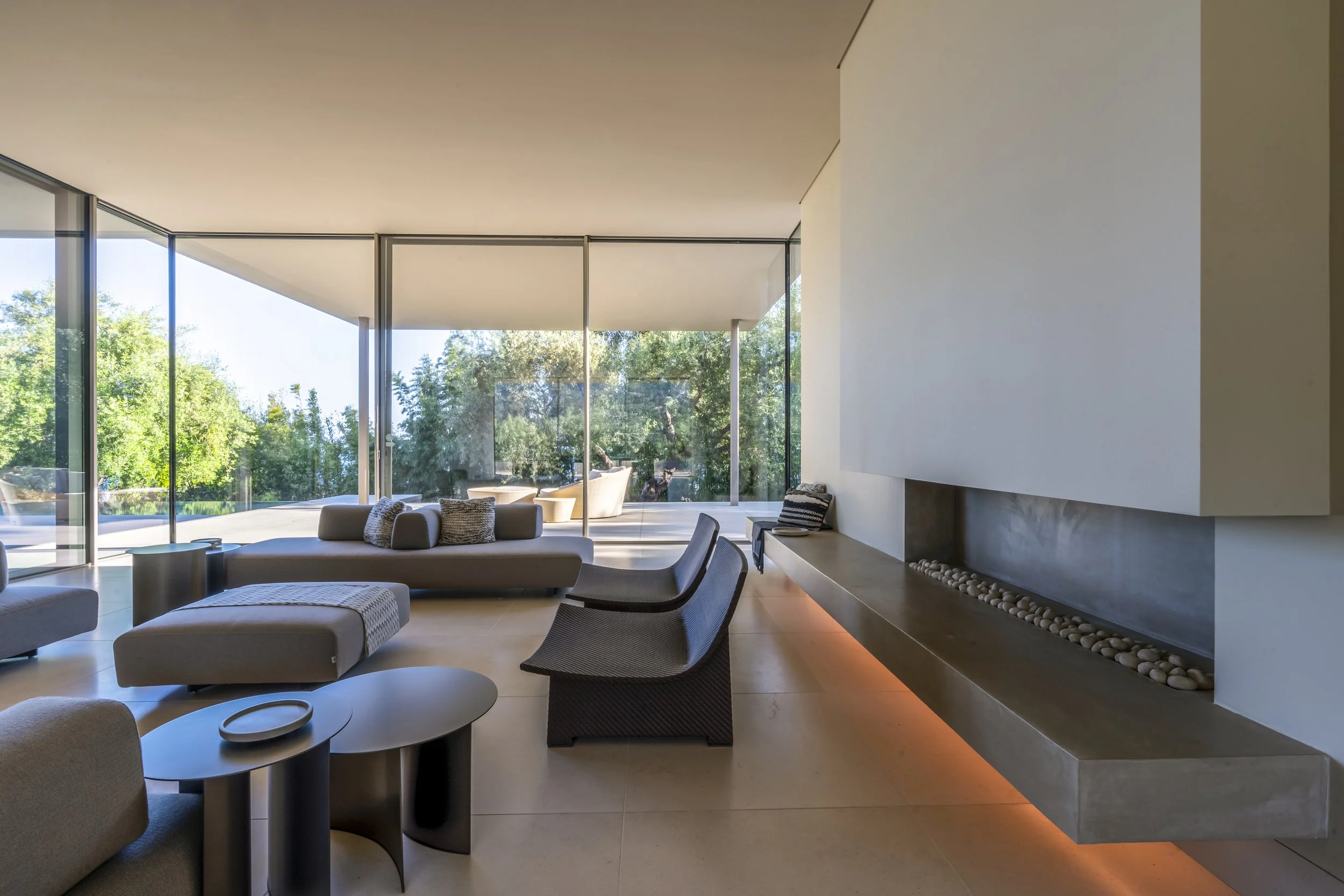 Modern living room at Toro Canyon Residence by The Warner Group Architects, Inc., featuring sleek furnishings, a minimalist fireplace, and floor-to-ceiling glass walls offering views of the surrounding natural landscape.