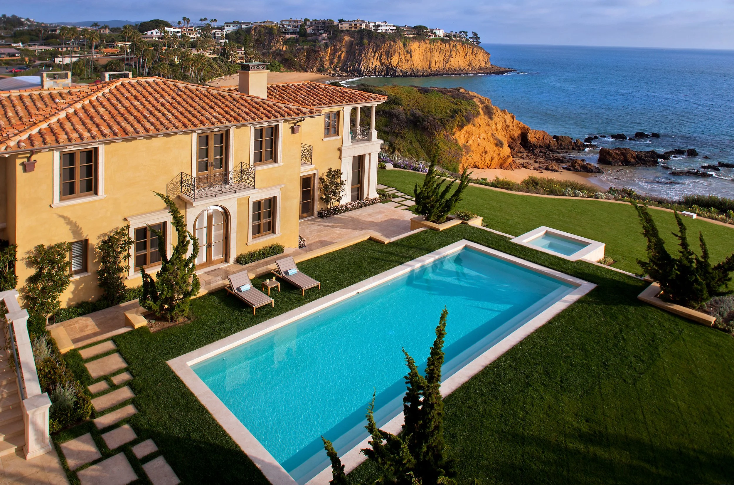 Luxury coastal home with a swimming pool overlooking the ocean, surrounded by green lawn and rocky cliffs.