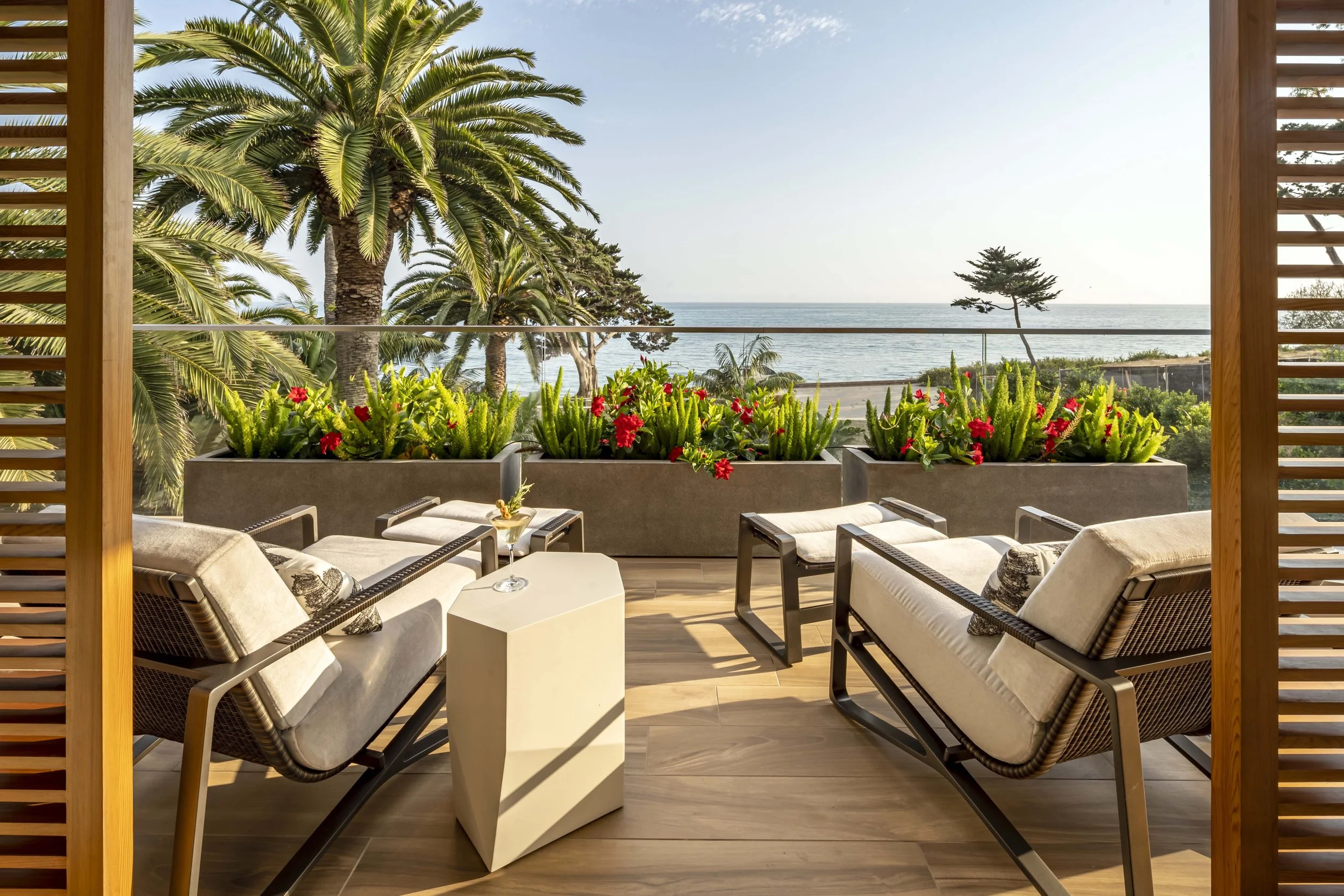 Outdoor patio with lounge chairs, small table, palm trees, ocean view, and vibrant potted plants.