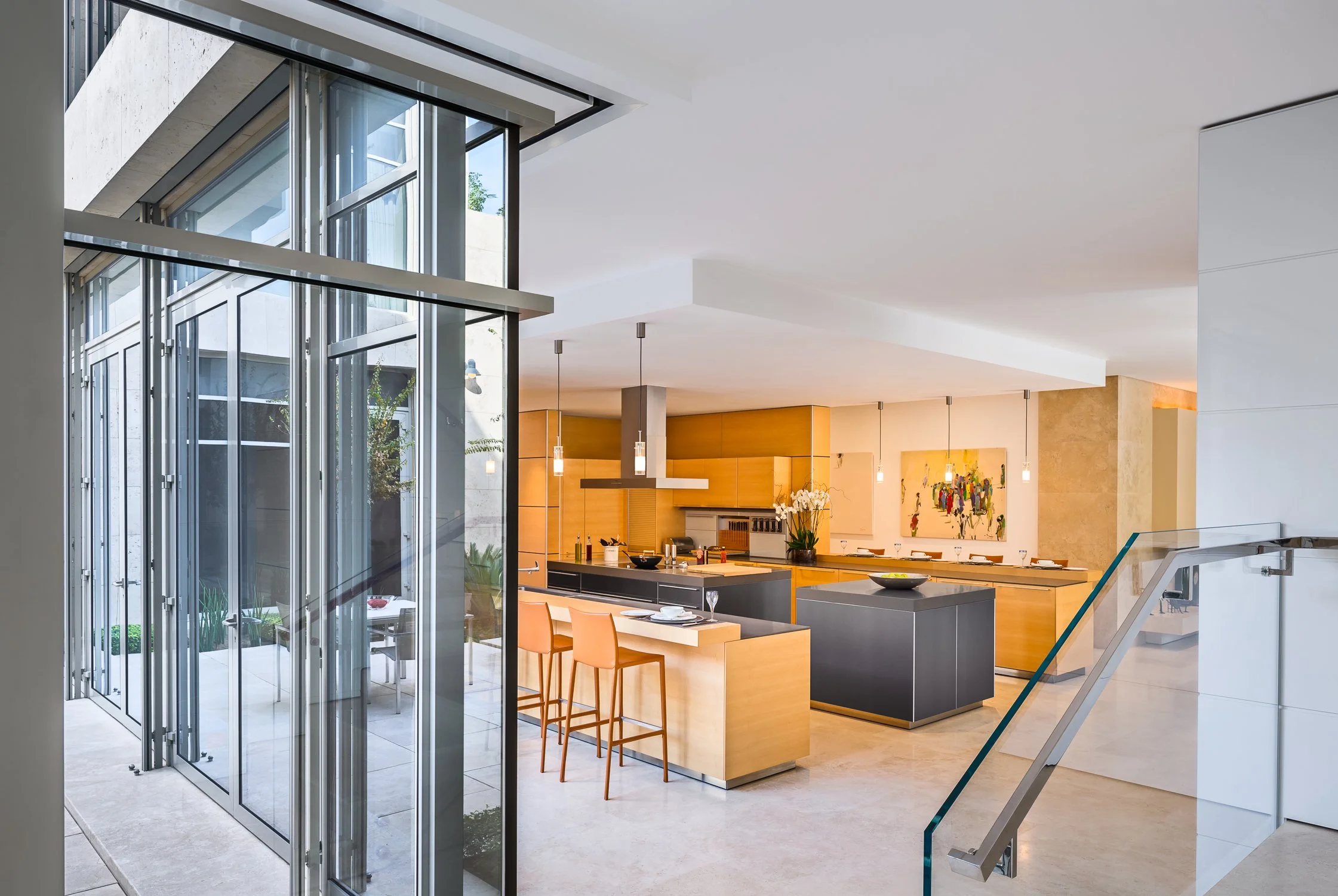 Modern kitchen with wooden cabinets, island, and bar stools, alongside glass windows and doors; pendant lights hang over the counter.