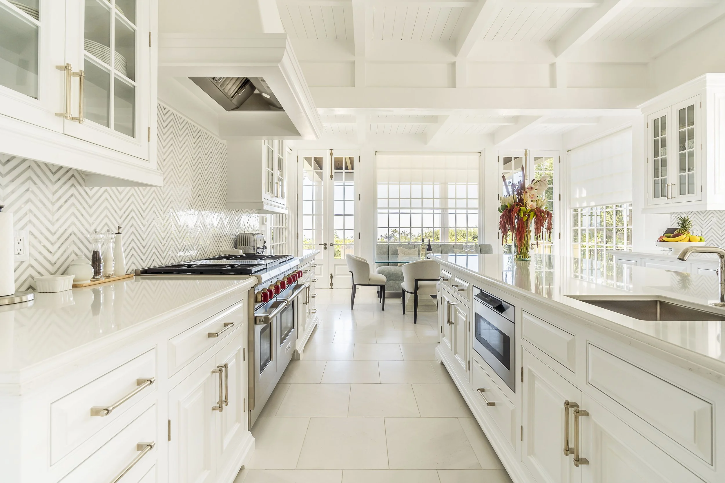 Spacious modern kitchen with white cabinetry, large island, and chevron backsplash, featuring stainless steel appliances and a breakfast nook, designed by The Warner Group Architects, Inc.