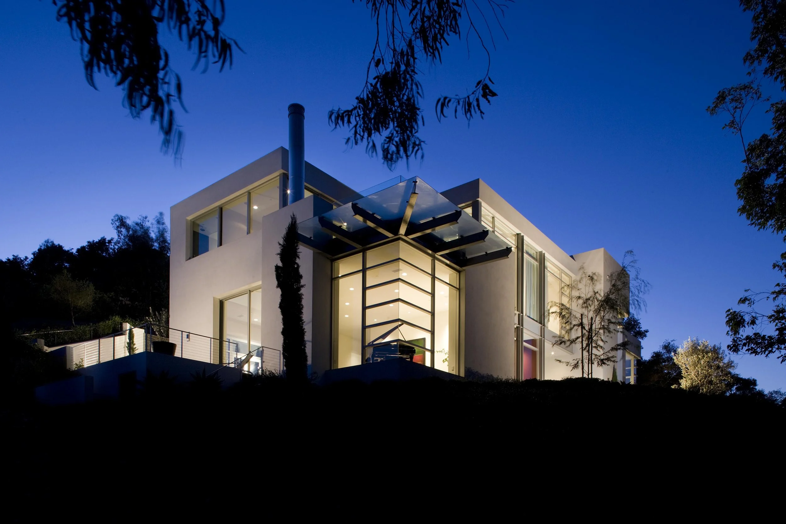 Modern hillside home illuminated at night with large glass windows and sleek architectural design, blending contemporary style with natural surroundings.